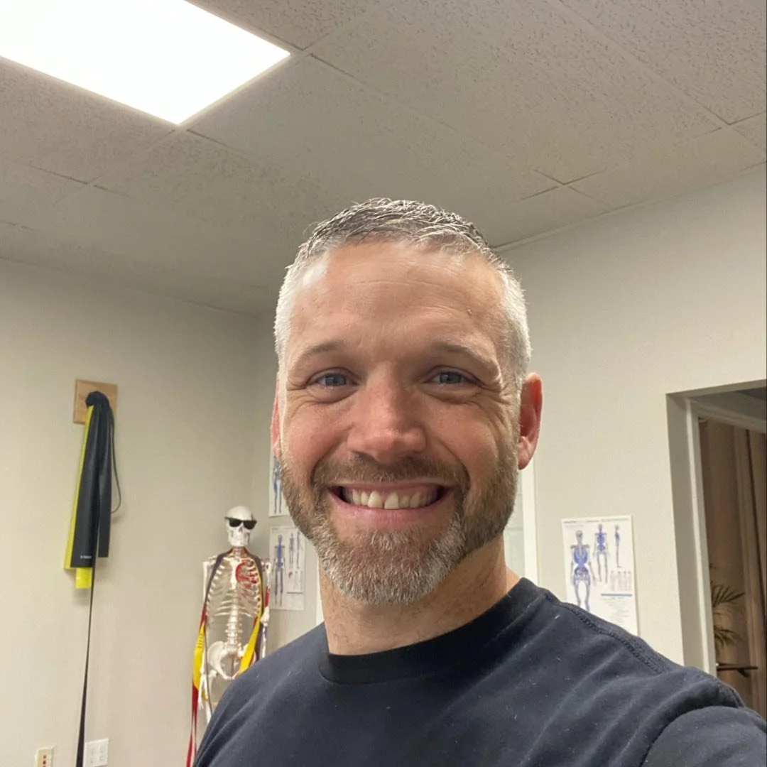 Smiling man with short gray hair and beard taking a selfie in a medical or physical therapy office, with anatomical posters and a skeleton model in the background.