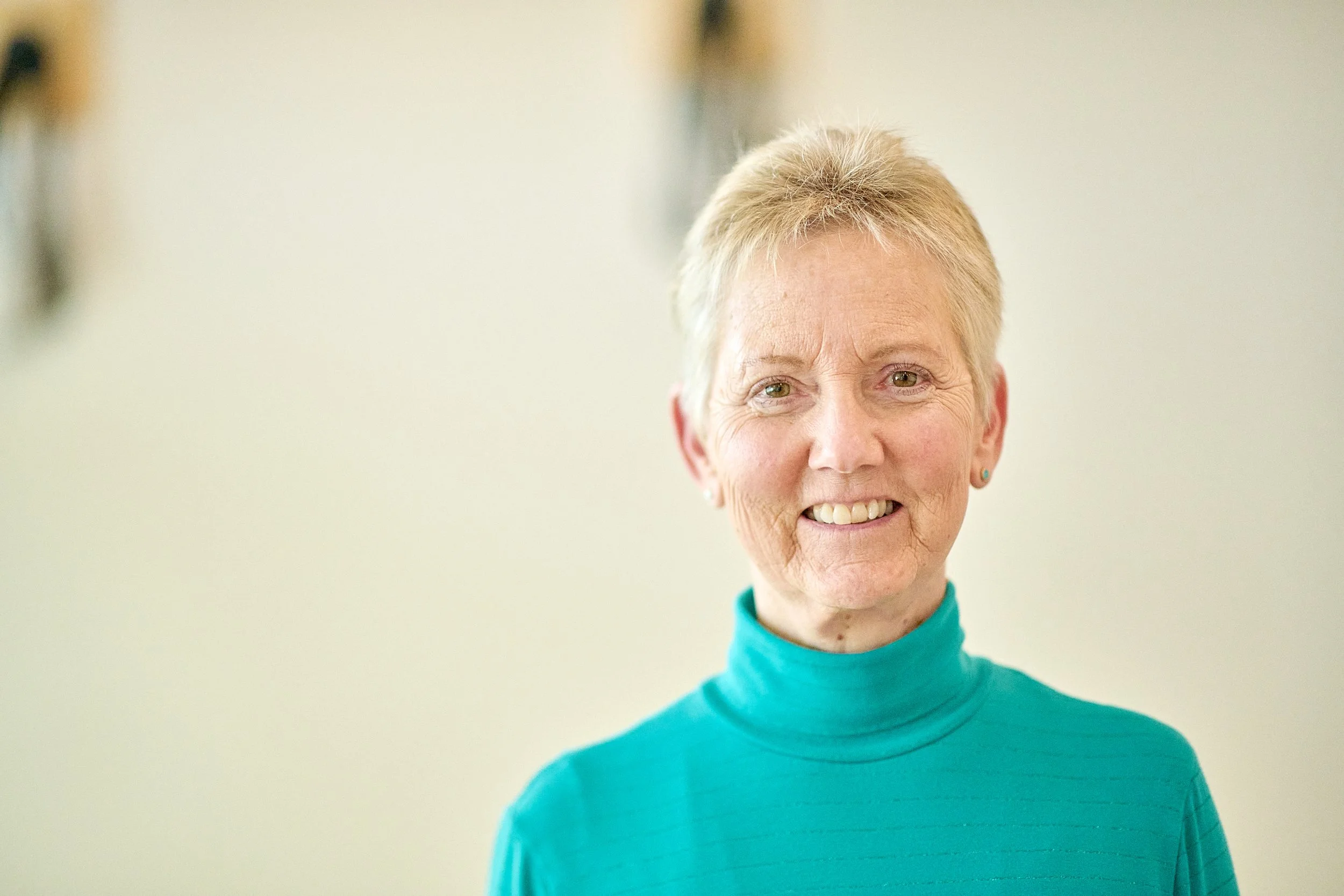 An elderly woman with short blonde hair and light skin, smiling and wearing a teal turtleneck shirt, standing against a plain background.