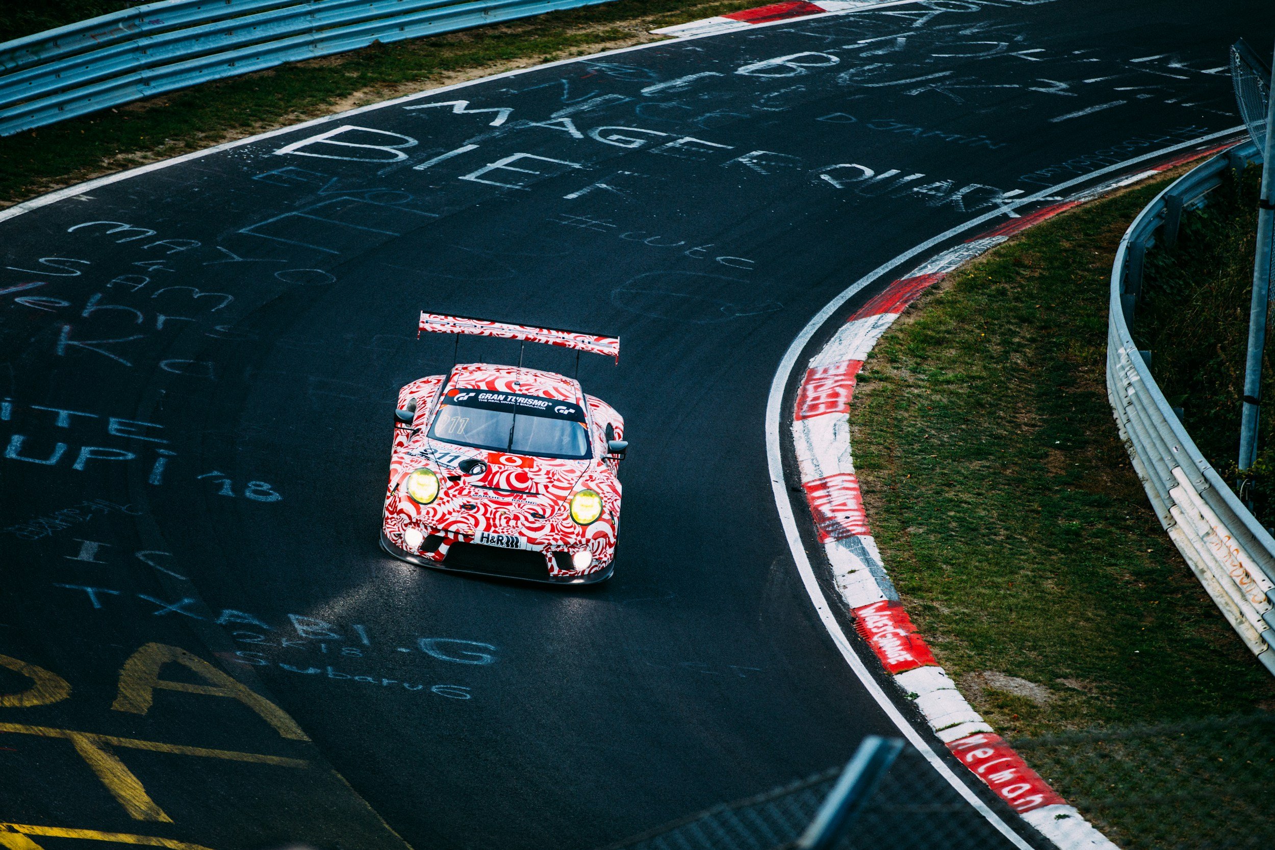A race car with a red and white swirl pattern on a wet race track with graffiti, turning a corner. The car has a large rear wing and yellow headlights.