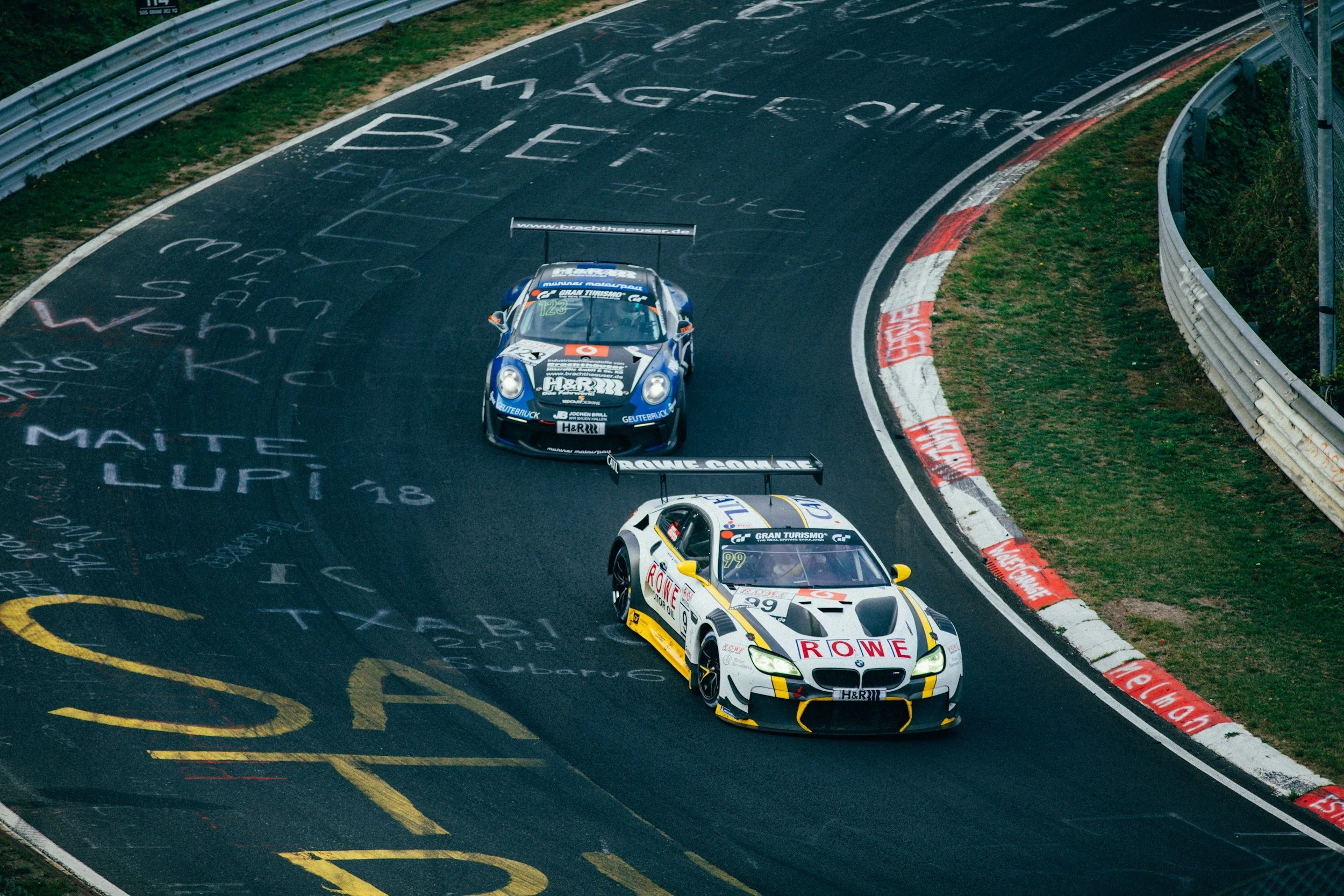Two race cars on a curving racing track with graffiti and messages written on it. The leading car is a white and black BMW with red and yellow accents, and the car behind is blue with white and orange accents.