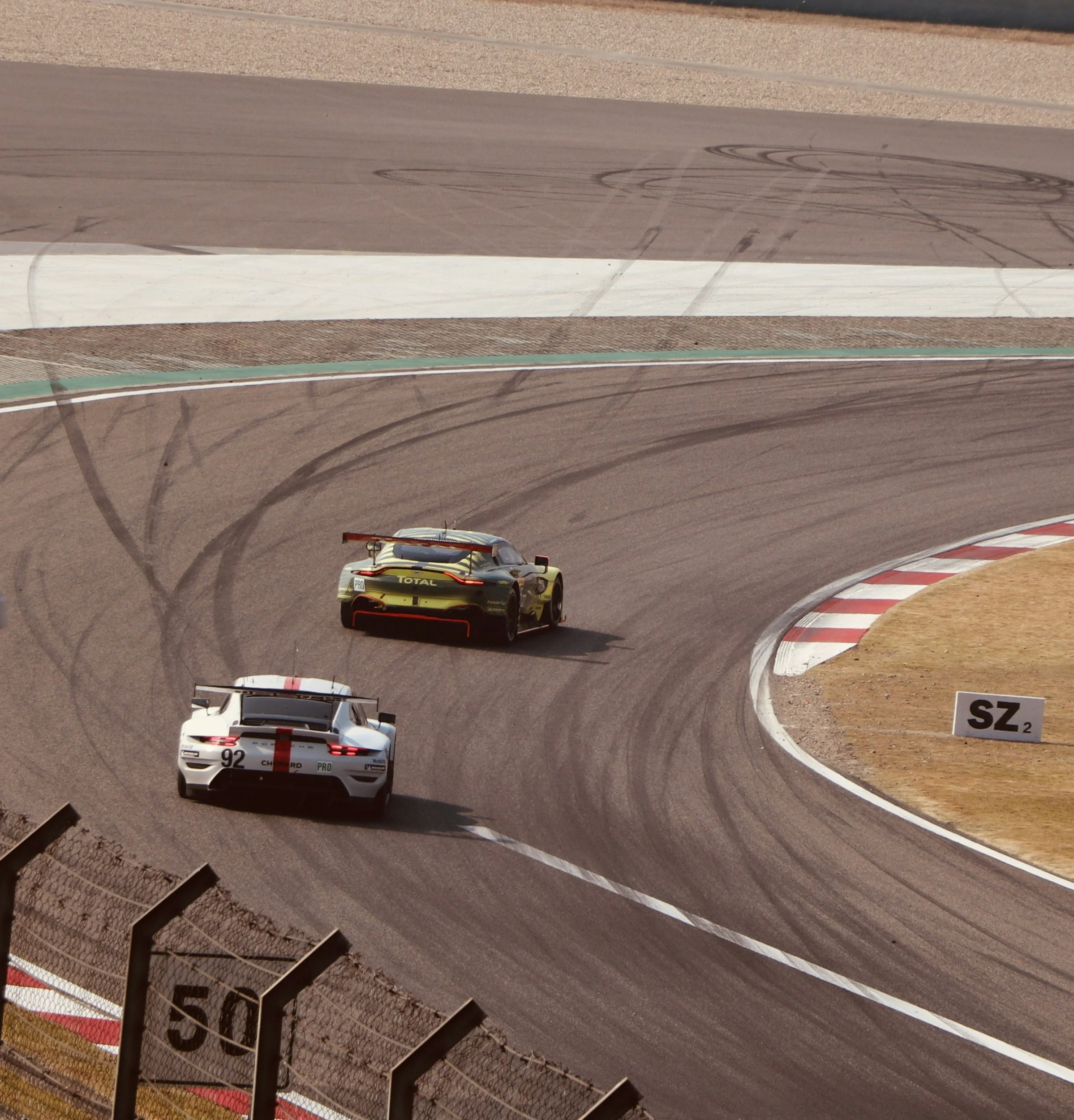 Two race cars on a curved asphalt track, one white with red markings and the other yellow with black accents, leaving tire marks as they navigate the turn.
