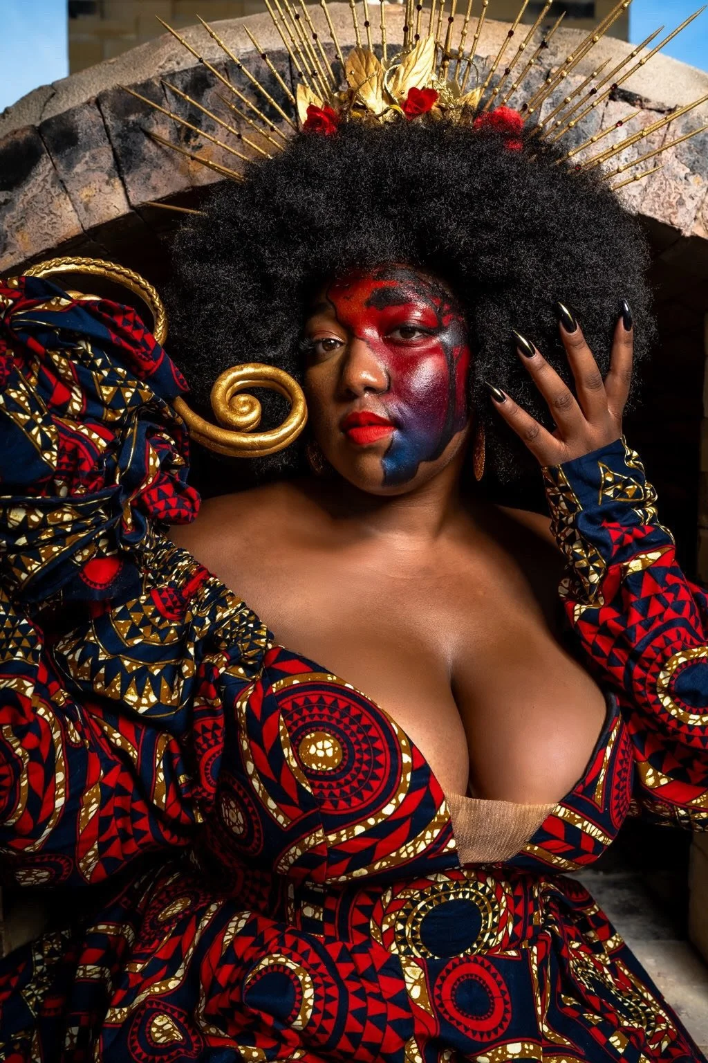 A woman dressed in vibrant African print fabric, with elaborate makeup and accessories, wearing a large Afro wig and gold jewelry, posing with a confident expression.