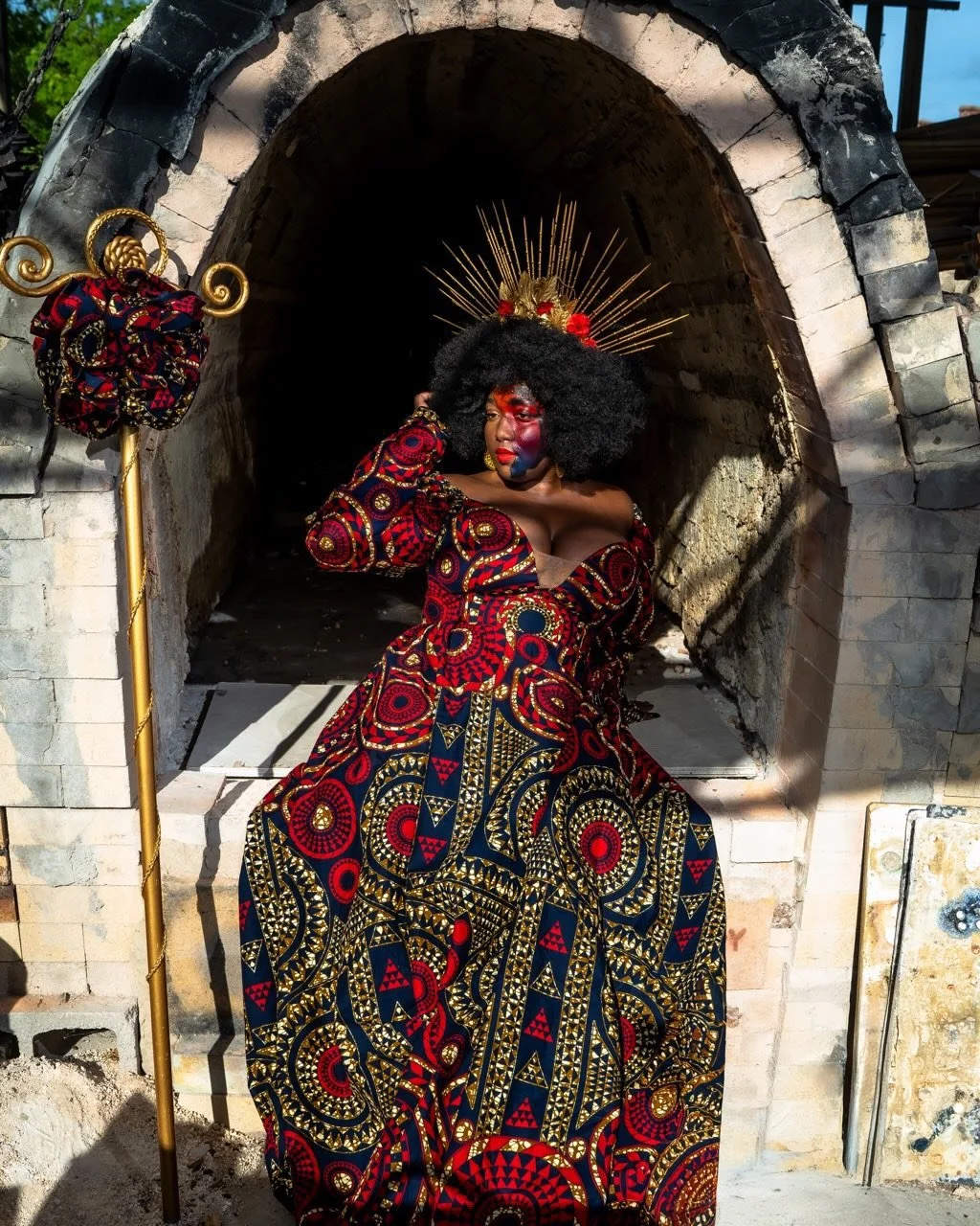 A woman dressed in a colorful, patterned African-style dress, wearing a black curly wig and a gold sunburst headpiece with red accents, standing in front of a brick oven or fireplace with a dark interior. She has vibrant red and blue face paint and is holding her head with one hand. A golden staff with a fabric-covered ornament on top stands beside her.