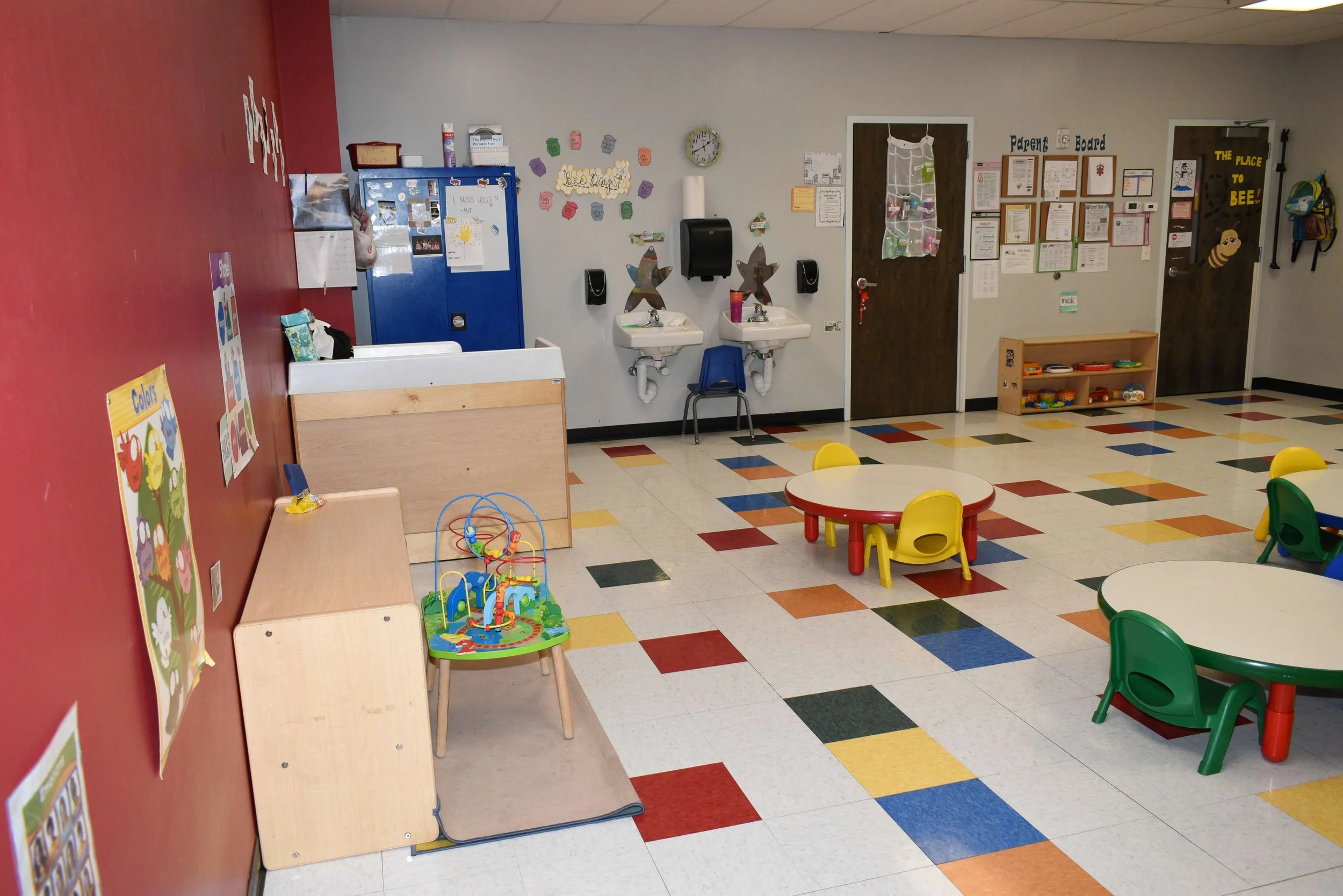 Empty preschool classroom with colorful tables and chairs, toy and play area, sinks, and bulletin boards on the walls.