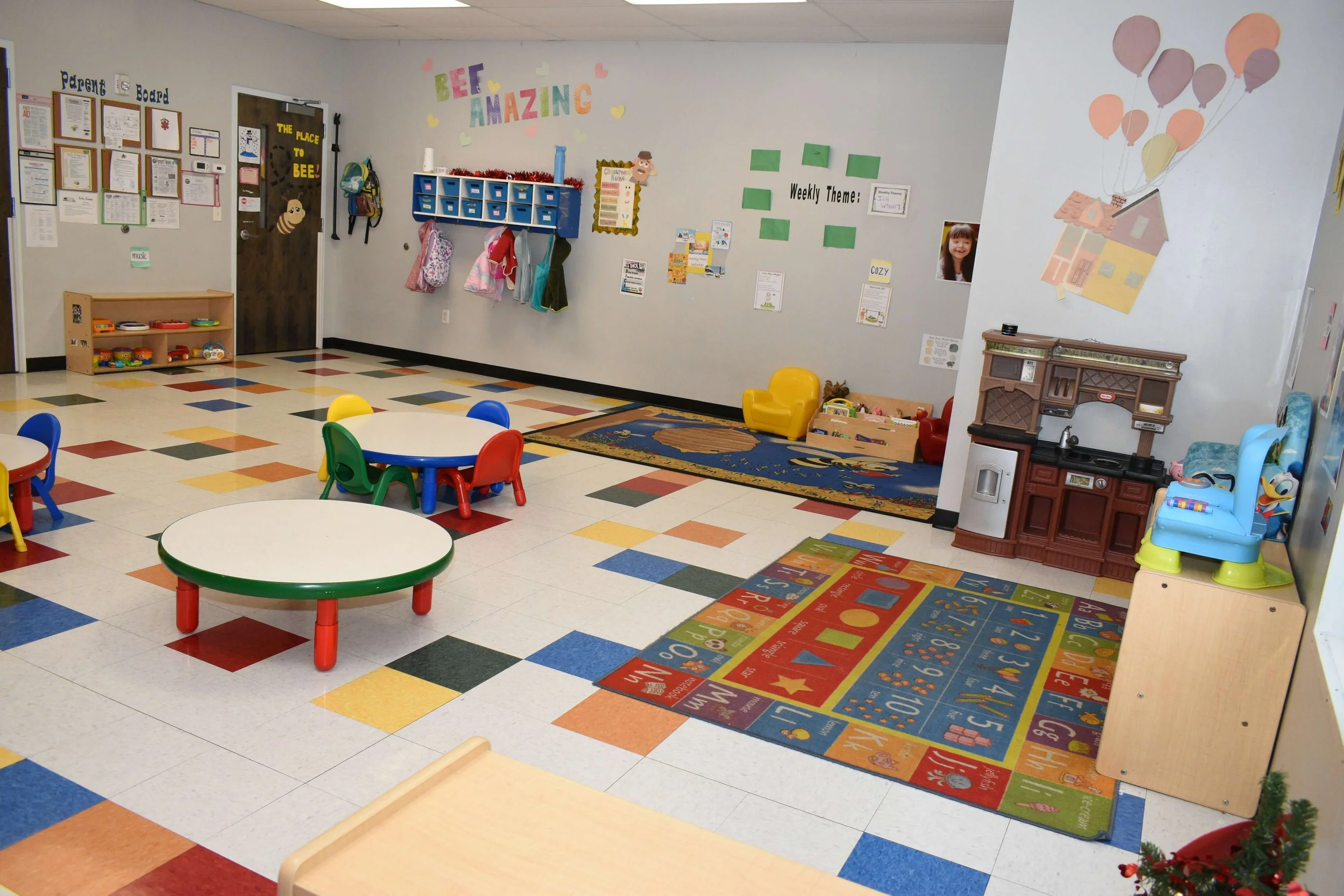 Colorful preschool classroom with small tables and chairs, toy kitchen set, wall decorations, and a play area with toys and a rug.