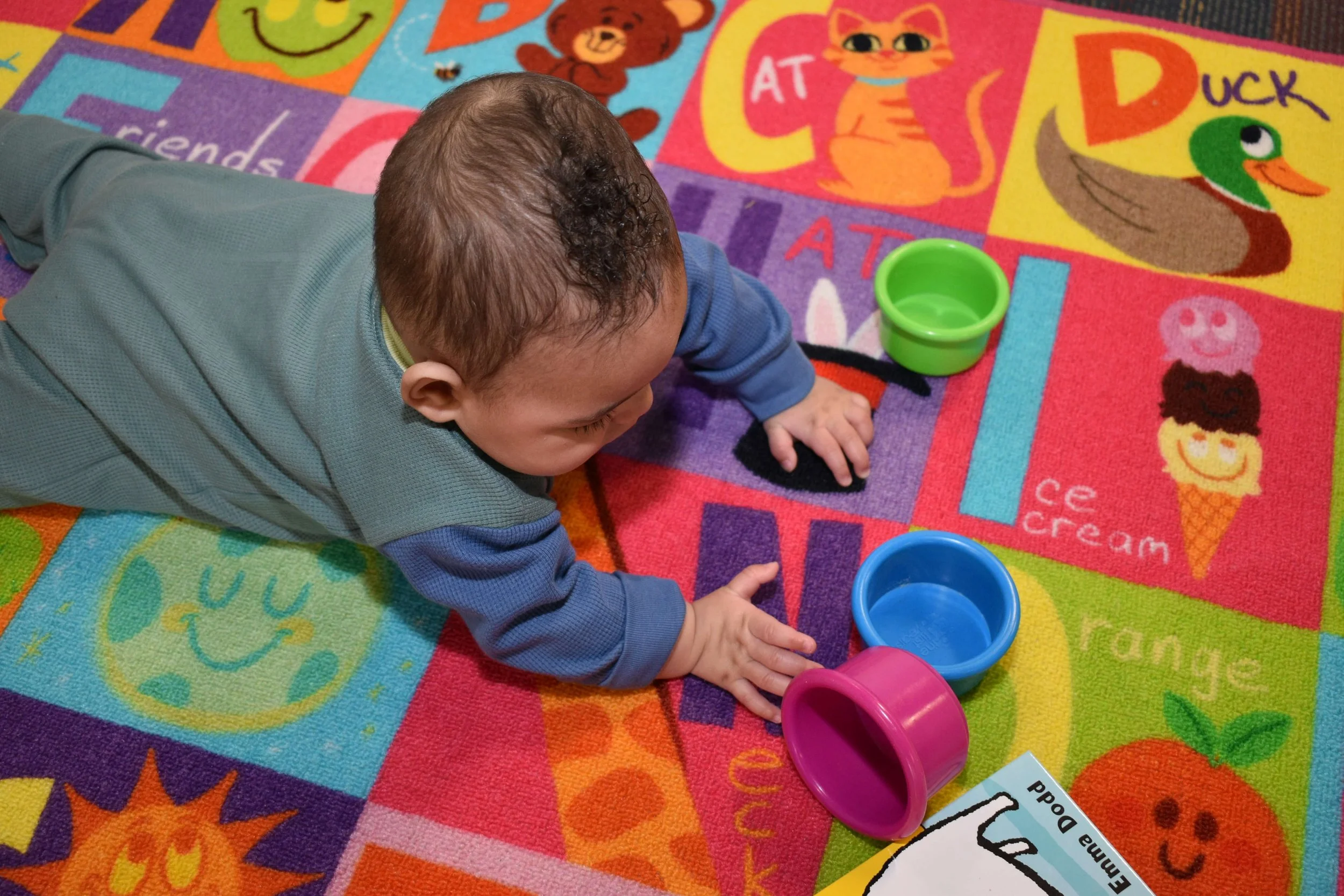 A young child lying on a colorful children's alphabet rug, engaging with plastic cups in green, blue, and pink, surrounded by educational illustrations and words.