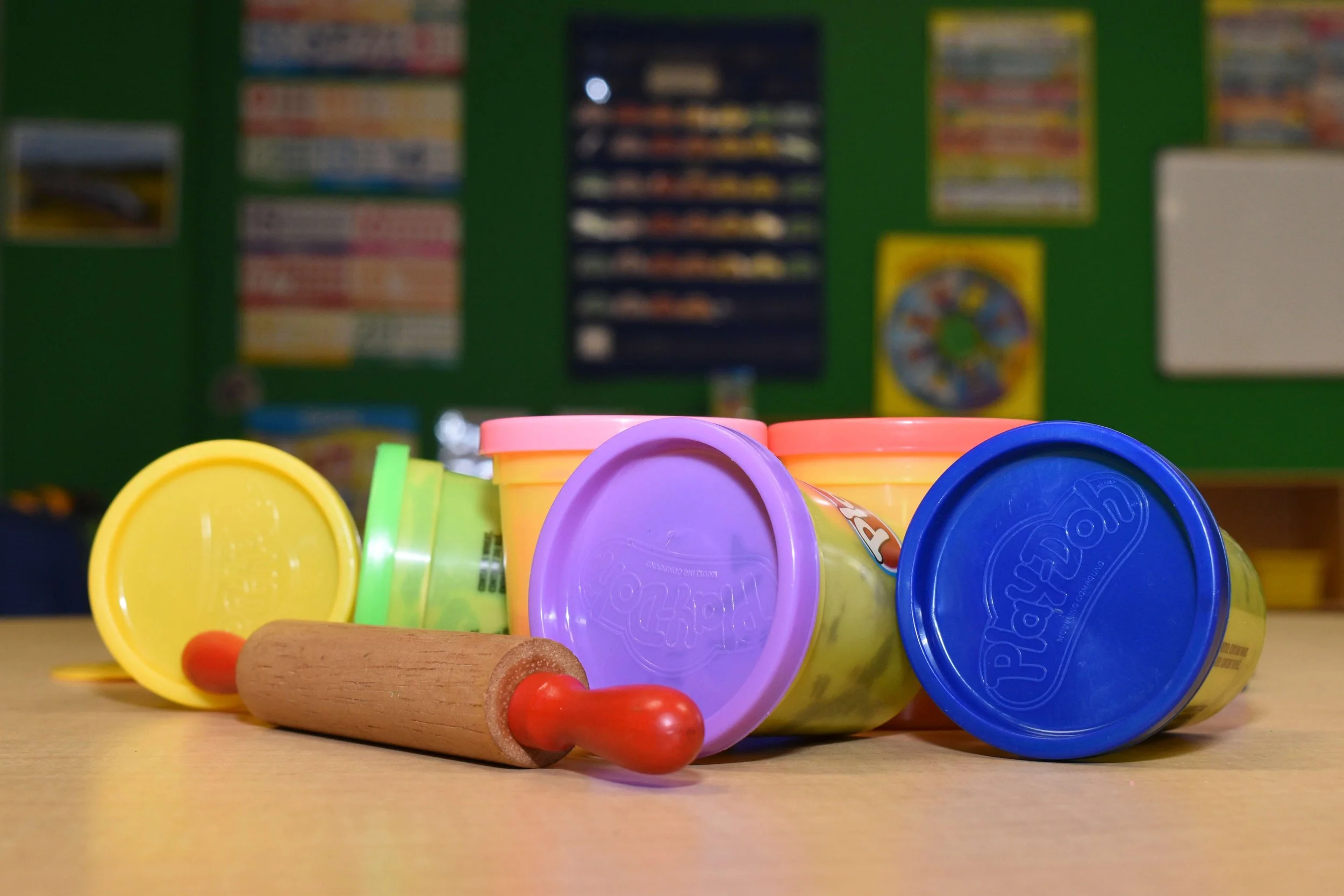 Colorful Play-Doh containers with a small rolling pin on a wooden surface, against a green classroom wall with posters and charts.