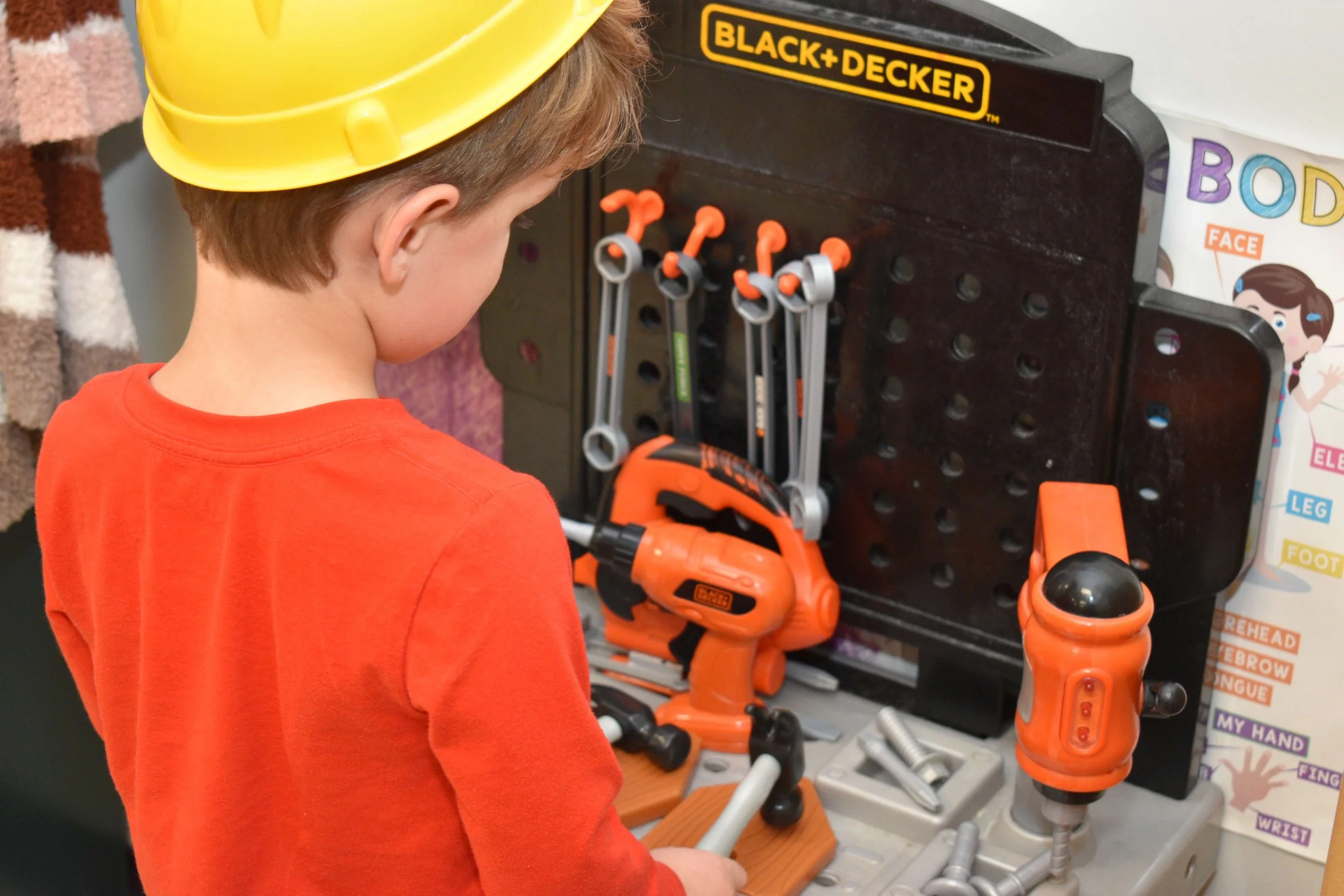 A young boy wearing a yellow hard hat and orange shirt playing with toy tools at a pretend workbench, which includes a toy drill, wrench, screwdriver, and other tools, set up in a classroom or play area.