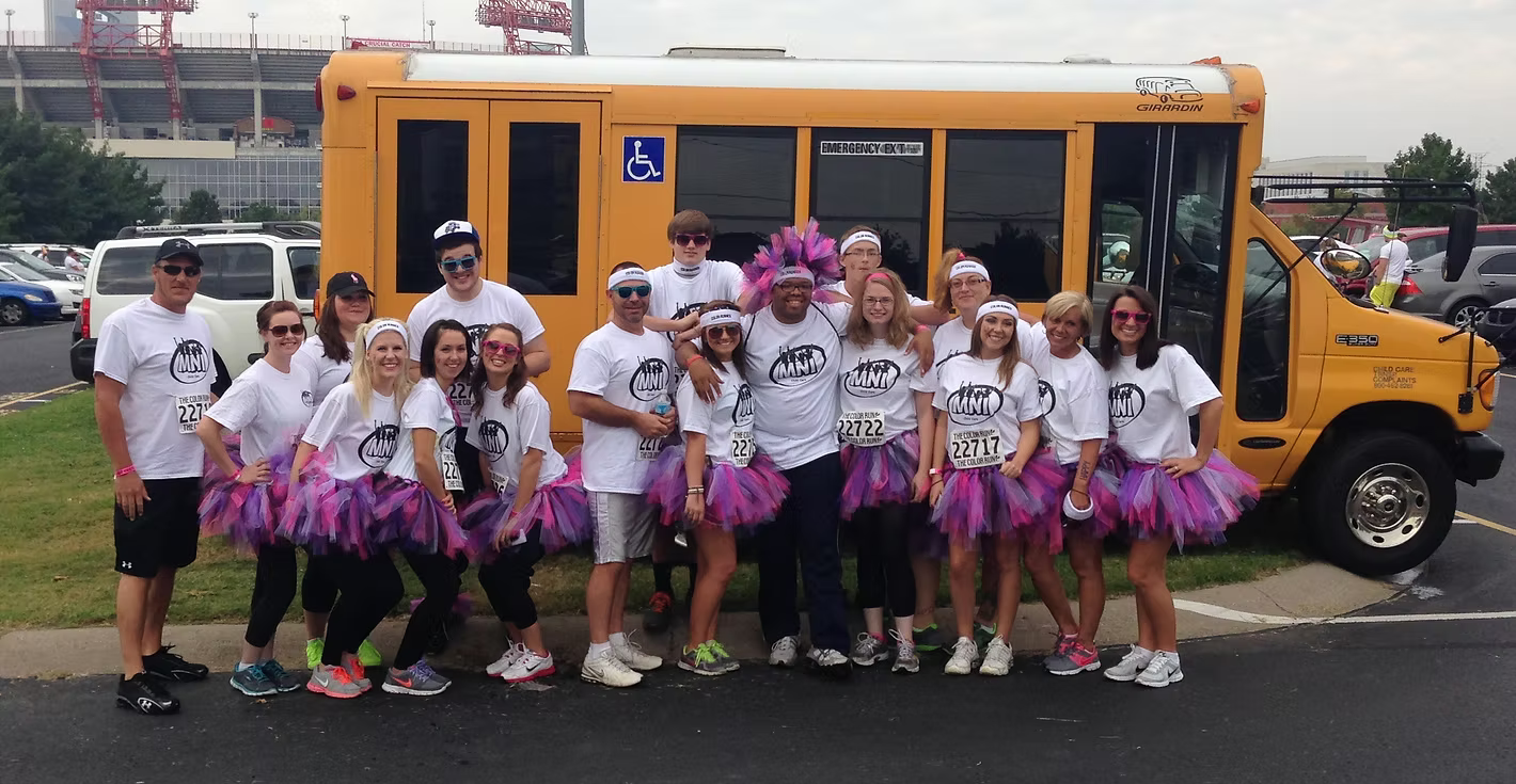 Group of MNI Kids staff members dressed in matching white T-shirts and colorful tutus posing in front of a school bus during the day, some wearing headbands and sunglasses, with a parking lot and a sports stadium in the background.