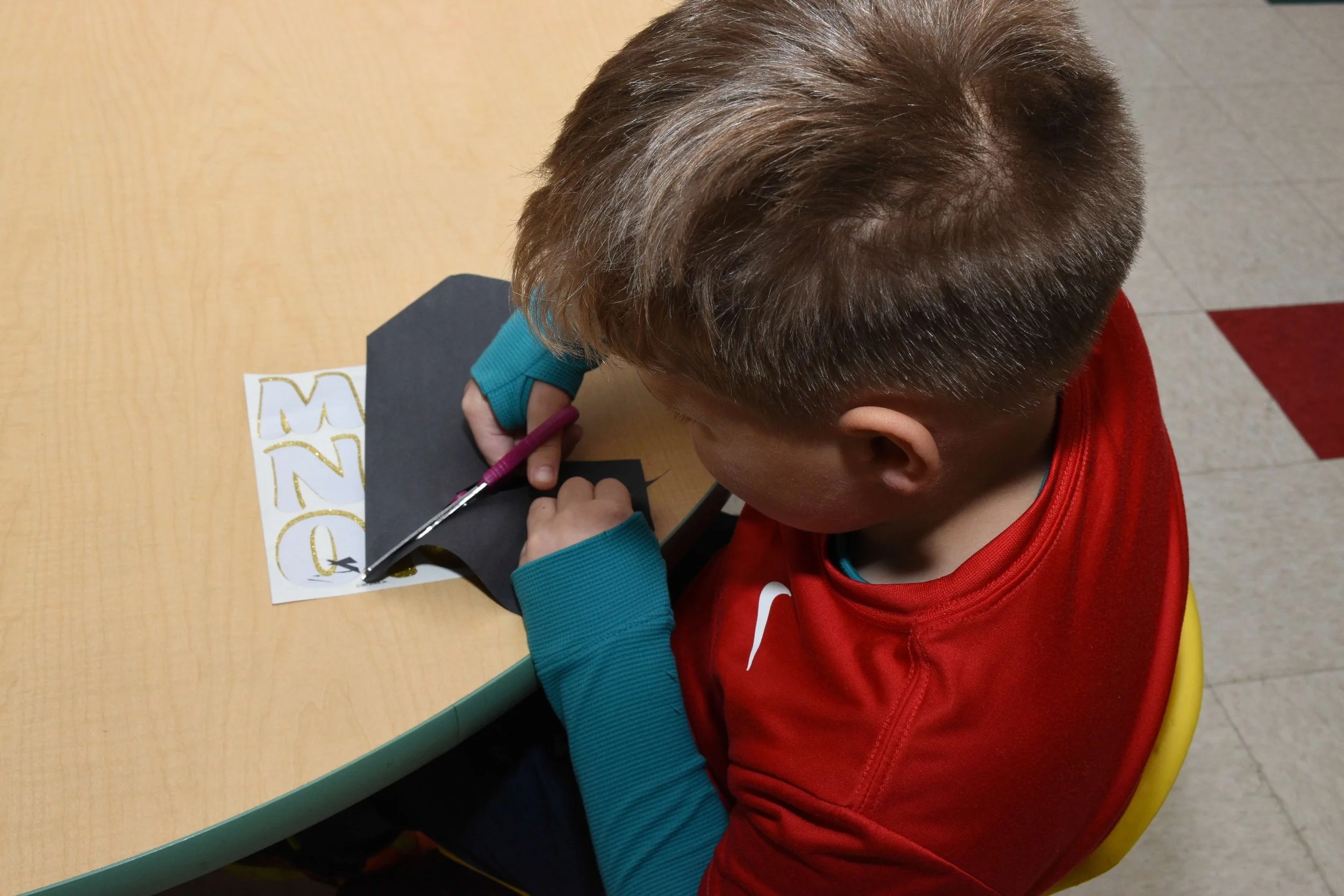 A young boy with short brown hair, wearing a red shirt and teal sleeves, is cutting a piece of black paper with pink scissors. There is a napkin with gold numbers 2022 on it on the table.