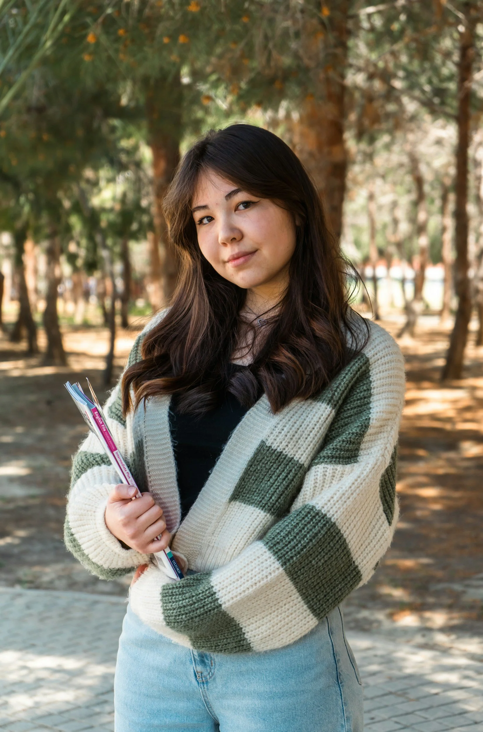 A young woman holding books and standing outdoors in a wooded area with trees in the background.
