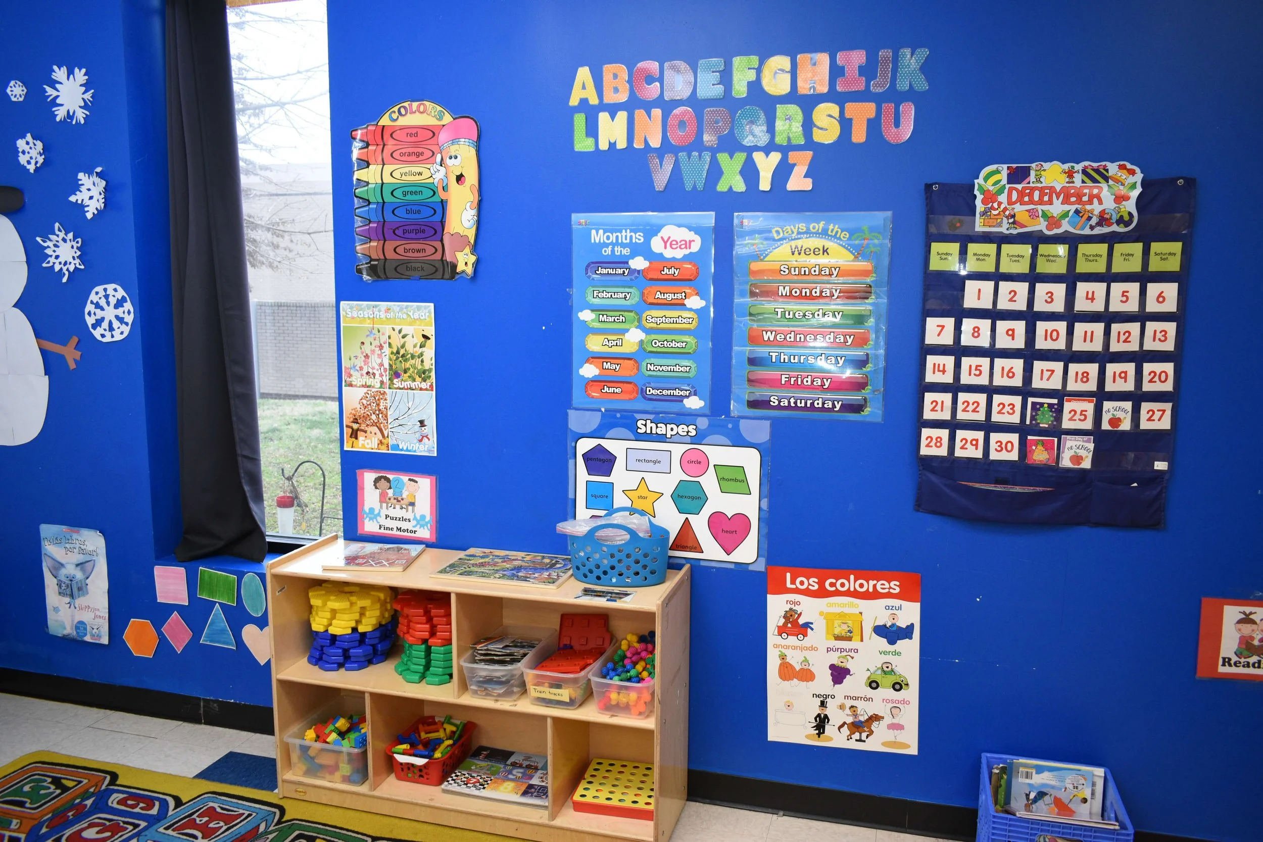 Colorful classroom wall with educational posters about the alphabet, months, seasons, shapes, days of the week, and numbers. There is a small wooden shelf with educational toys and books.