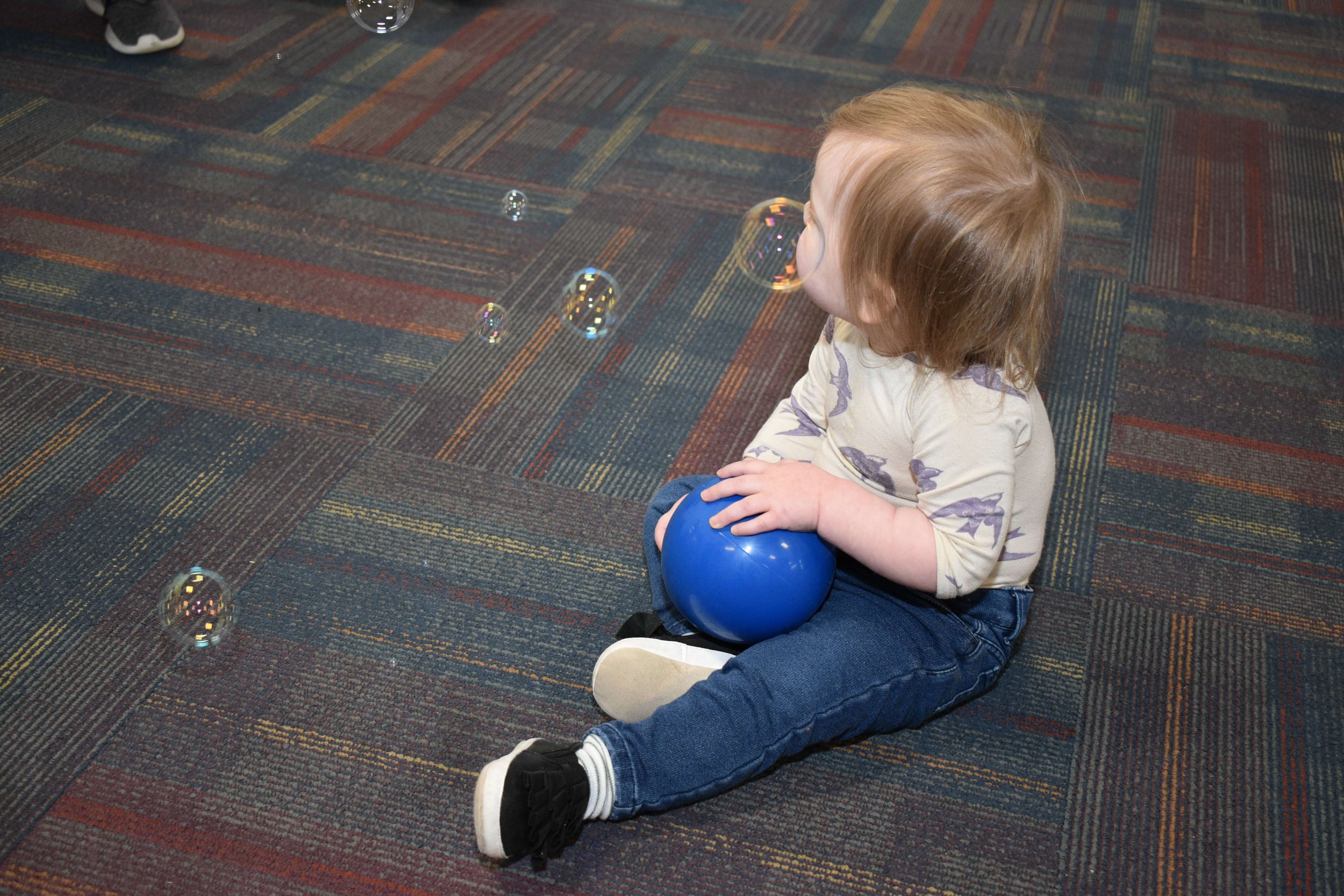 A young child with light brown hair sitting on a multicolored carpet, holding a blue ball and surrounded by soap bubbles.