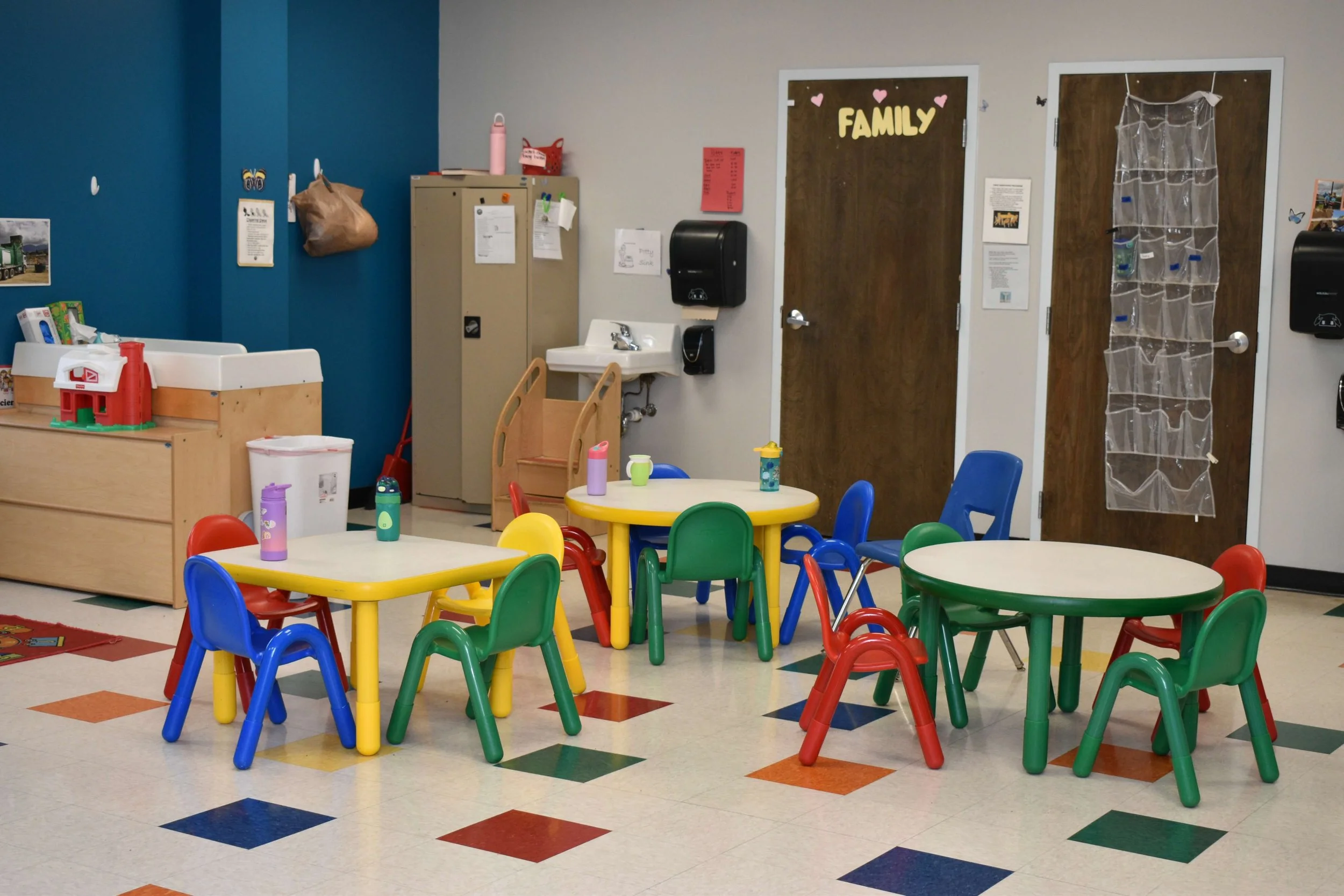 Colorful children's playroom with small round tables, green, blue, red, and yellow chairs, water bottles, a wooden changing table, a locker, a handwashing sink, and two wooden doors, one decorated with the word 'FAMILY' and pink hearts.