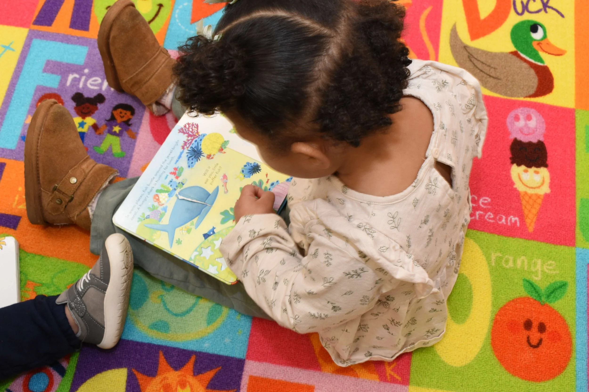 A young girl with dark, curly hair and light skin is sitting on a colorful educational carpet, reading a children's book about sea creatures, including a whale and a fish.
