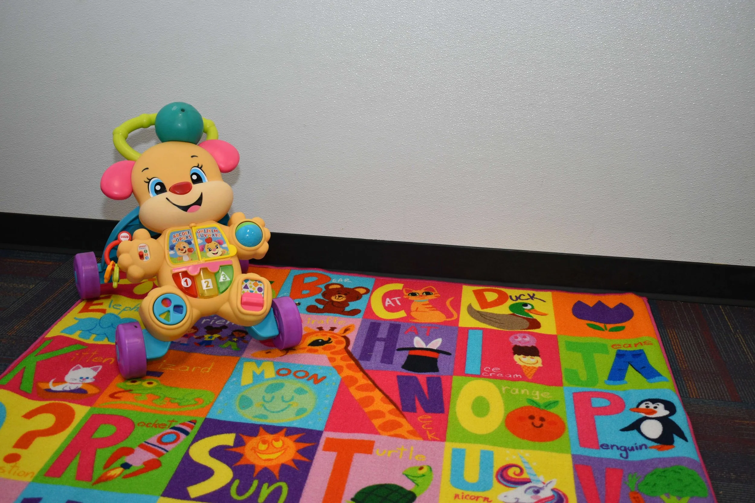 Colorful children's play area with a toy walker featuring a smiling cartoon animal face, placed on a vibrant alphabet-themed rug with illustrations of animals and objects.