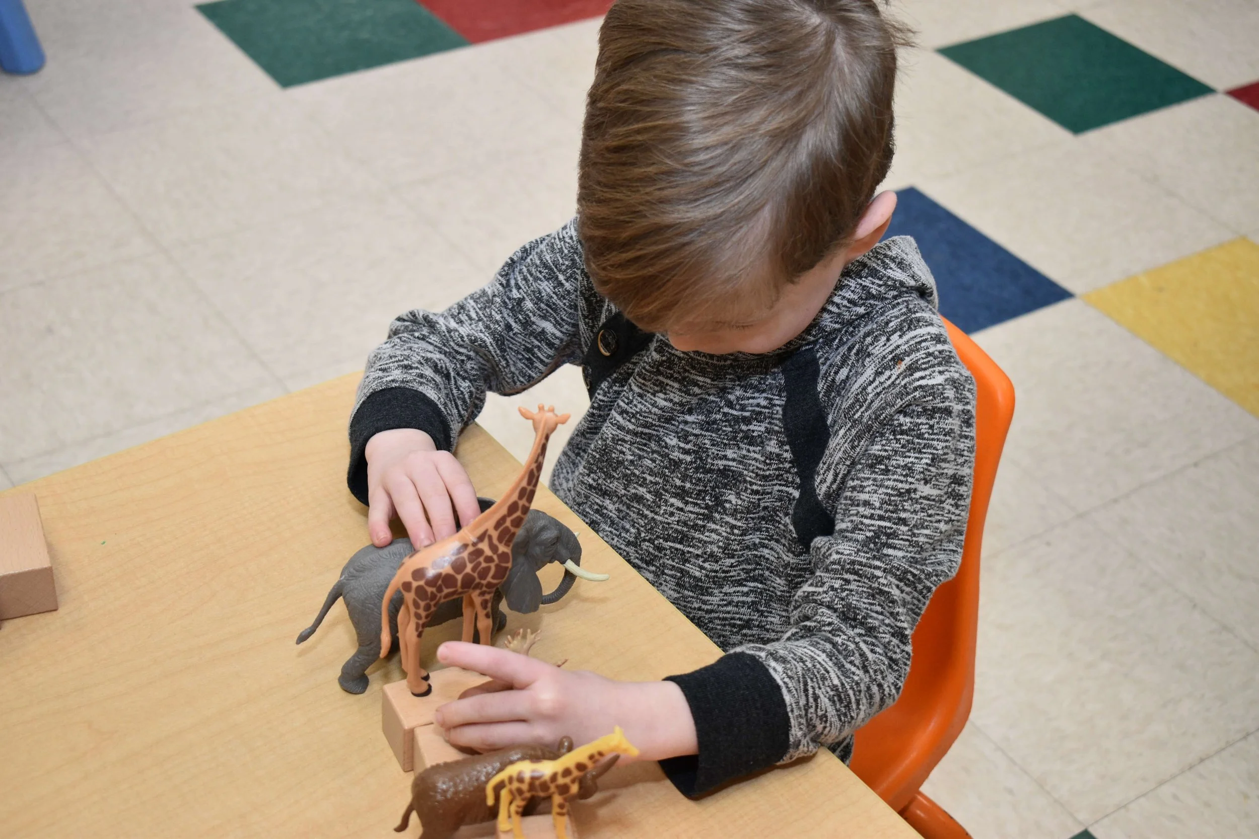 A young boy sitting at a wooden table playing with plastic toy animals, including a giraffe and an elephant in a room with a colorful tiled floor.