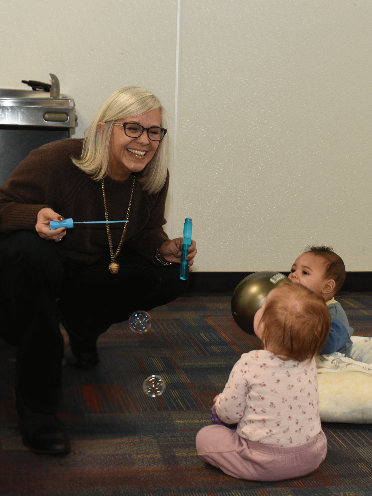 Nichole, President of MNI Kids, playing with two young children on the floor, blowing bubbles and holding a bubble wand, in an indoor setting with a carpeted floor and a countertop in the background.