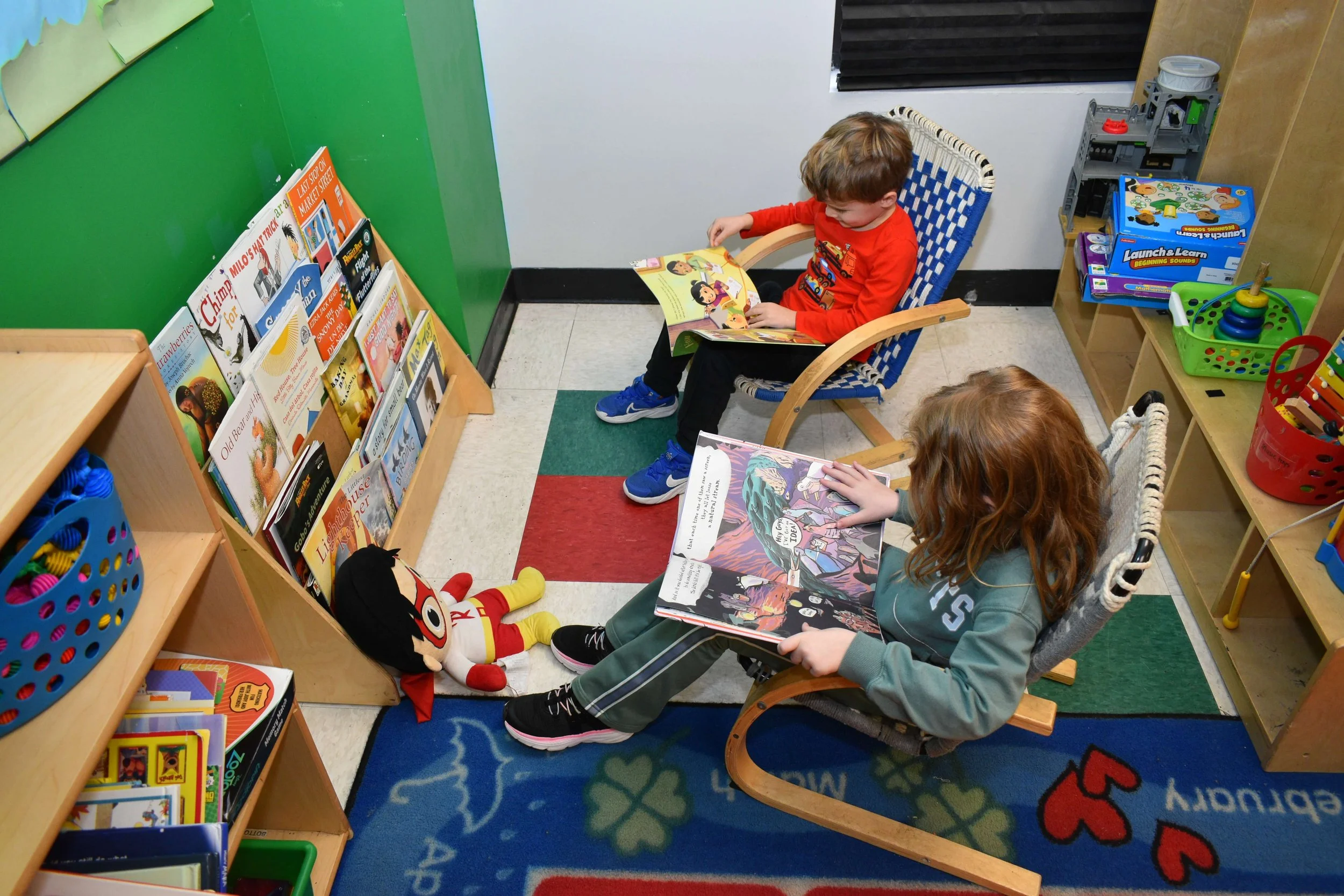 Two children reading picture books in a children's library corner with shelves of books and toys.