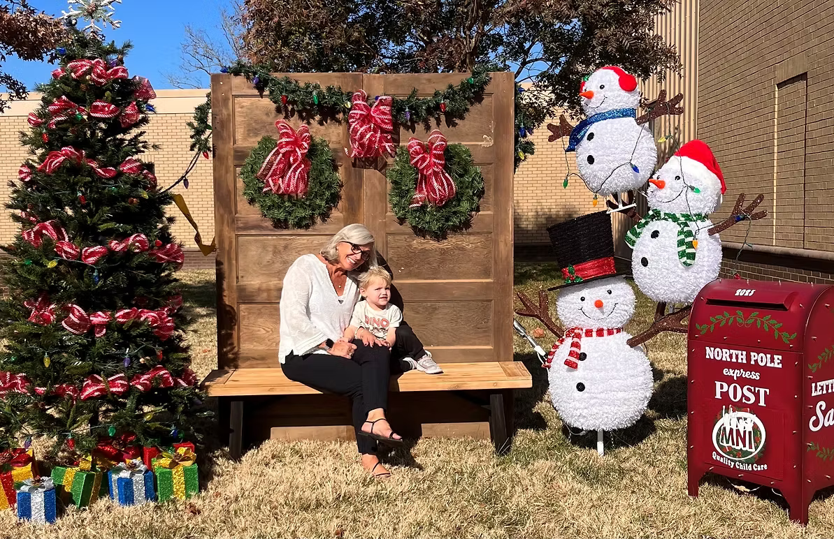 Nichole DePietro and a young boy sitting on a wooden bench, smiling in front of a Christmas-themed holiday display with decorated trees, wreaths, and snowman figures, outside on grass.