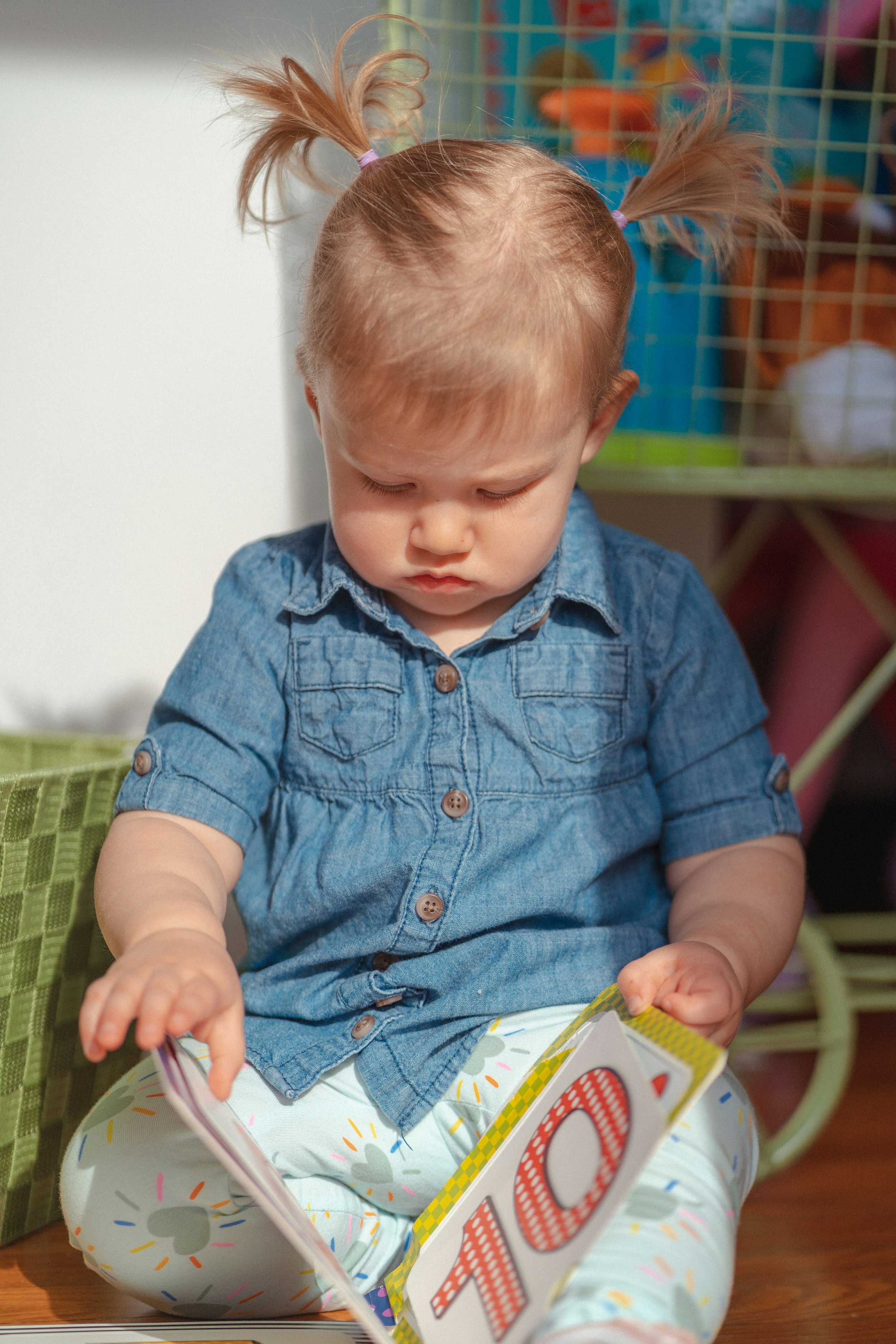 Young child with pigtails, wearing a denim shirt, sitting on the floor and looking at a colorful picture book.