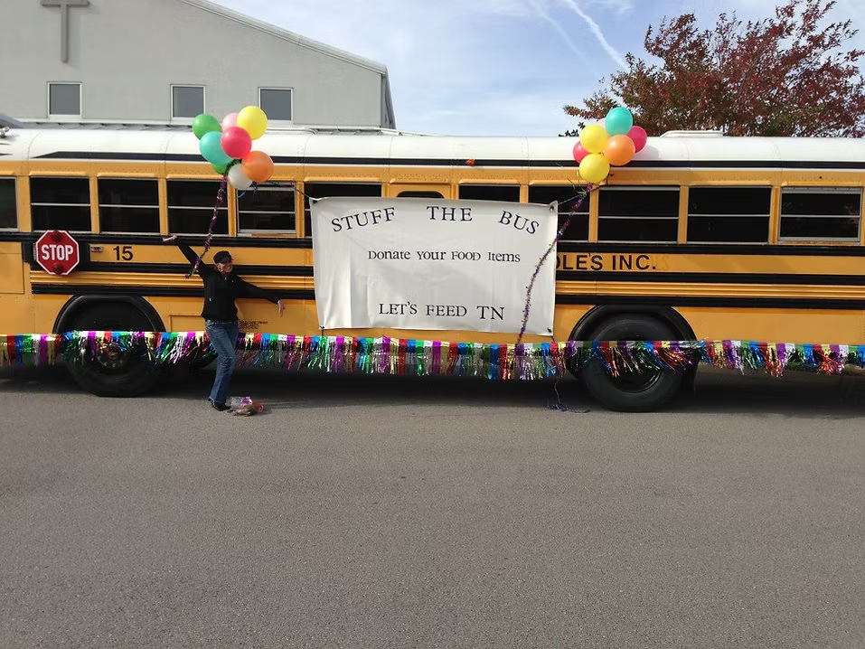 A decorated yellow school bus with colorful balloons and streamers, and a sign that reads "Stuff the Bus, Donate your food items, Let's feed TN." A person is standing beside the bus holding a bunch of balloons.