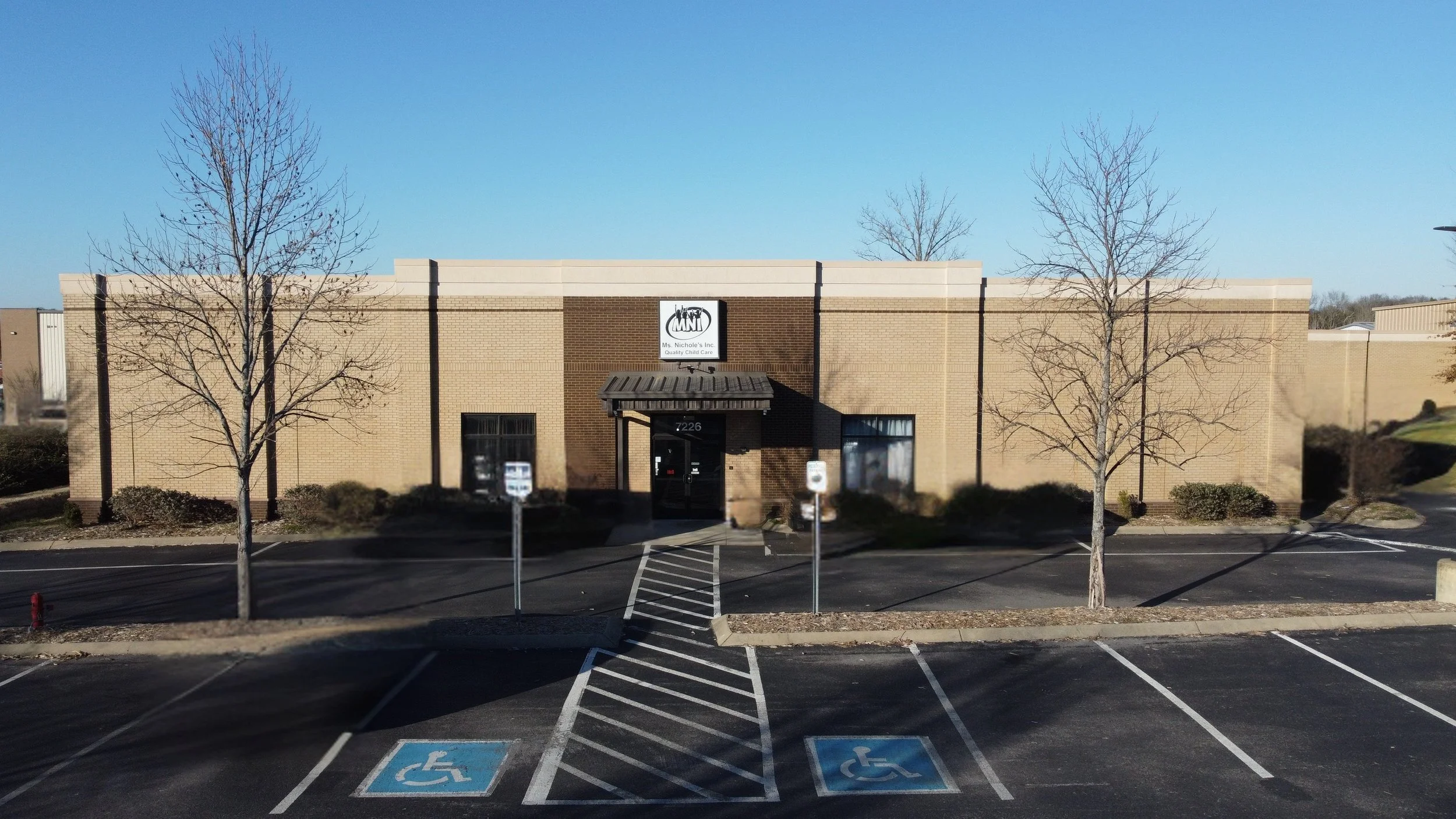 Exterior view of MNI Kids Childcare in Nolensville TN. The building has three trees in front, a parking lot with handicap parking spaces, and a clear blue sky.