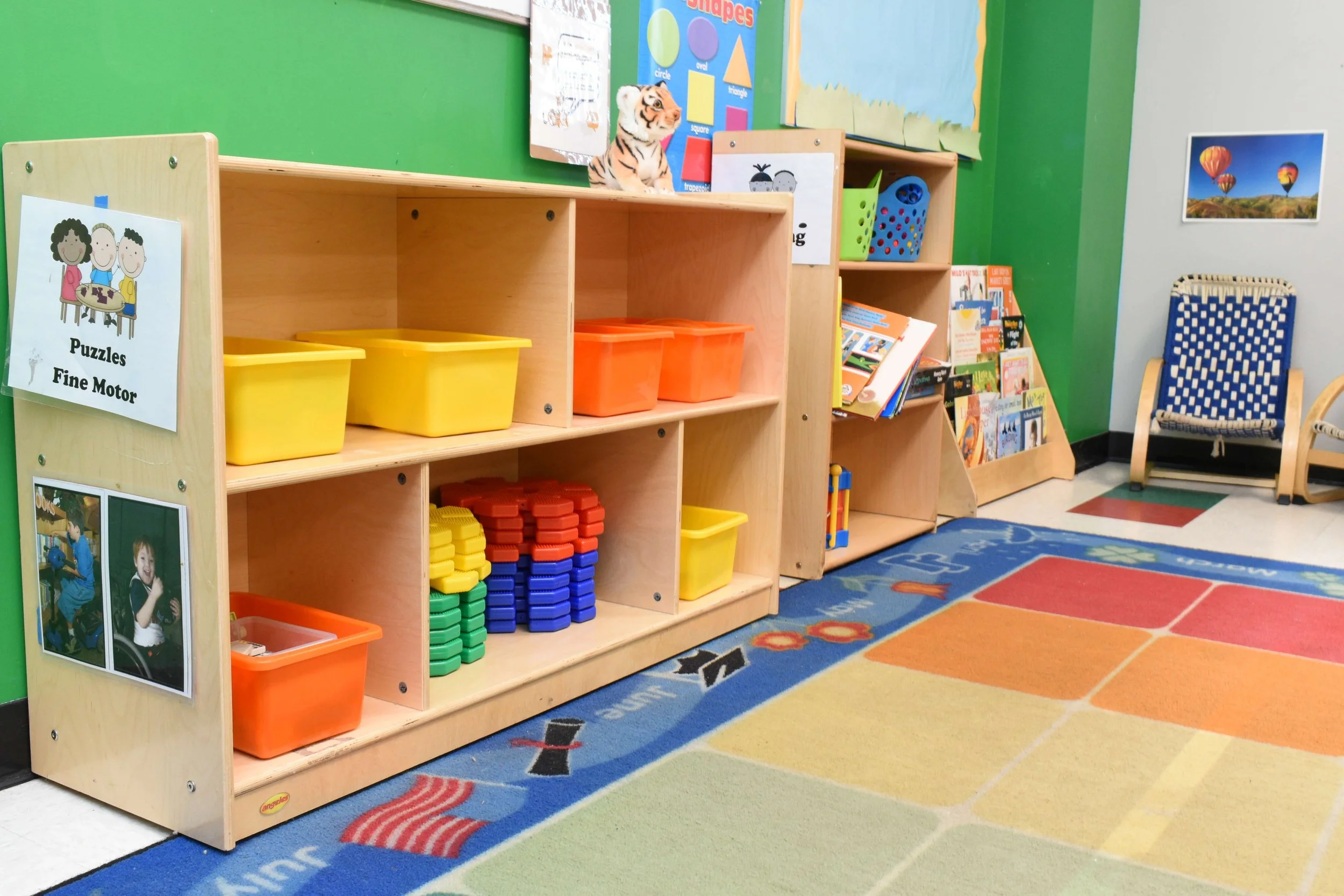 Classroom corner with wooden shelves containing yellow and orange storage bins, educational books, a poster about shapes, and a tiger plush toy on top. The floor has a colorful rug with squares and alphabet images. There are also a blue-and-white woven chair, wall art including hot air balloons, and a small bookshelf with more books.