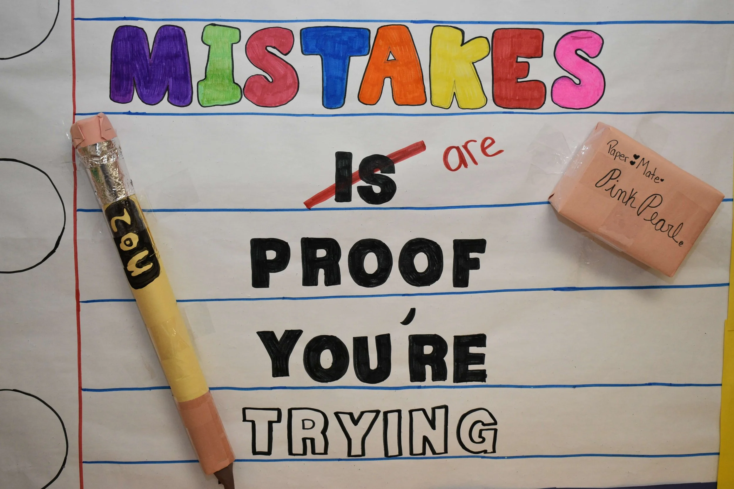 Colorful poster with the words 'Mistakes are proof you're trying' written on it. The word 'Mistakes' is in rainbow colors, and other words are in black, red, and outlined black fonts. There is a pink eraser labeled 'Pink Pearl' and a yellow pen with 