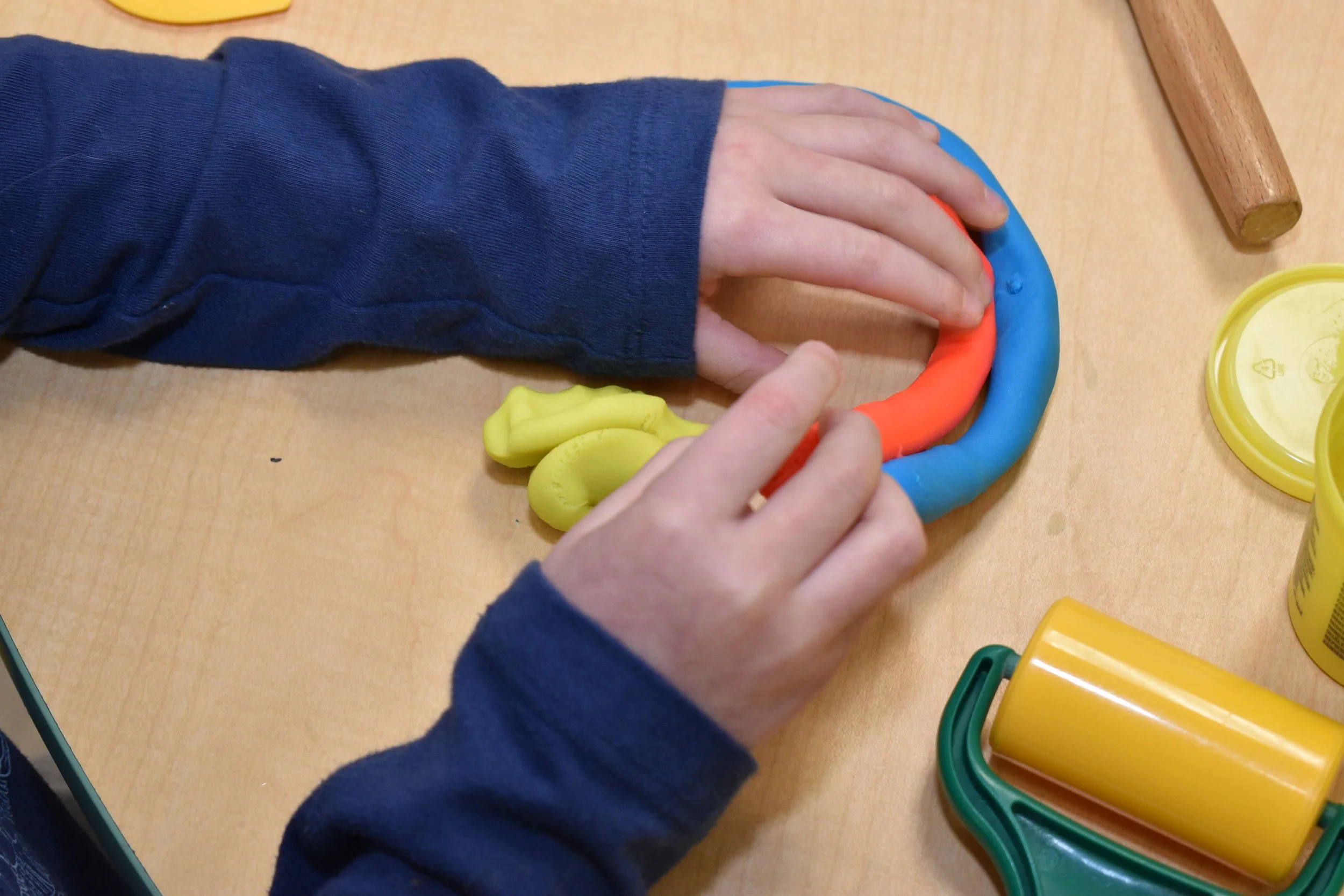 Hands of a child shaping colorful Play-Doh on a wooden table, with Play-Doh tools and containers nearby.