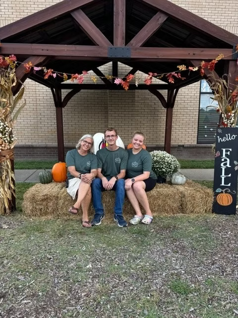 Three people sitting on hay bales under a wooden pavilion decorated with fall-themed garland, pumpkins, and hay bobs, in front of a building with a brick wall and a window.