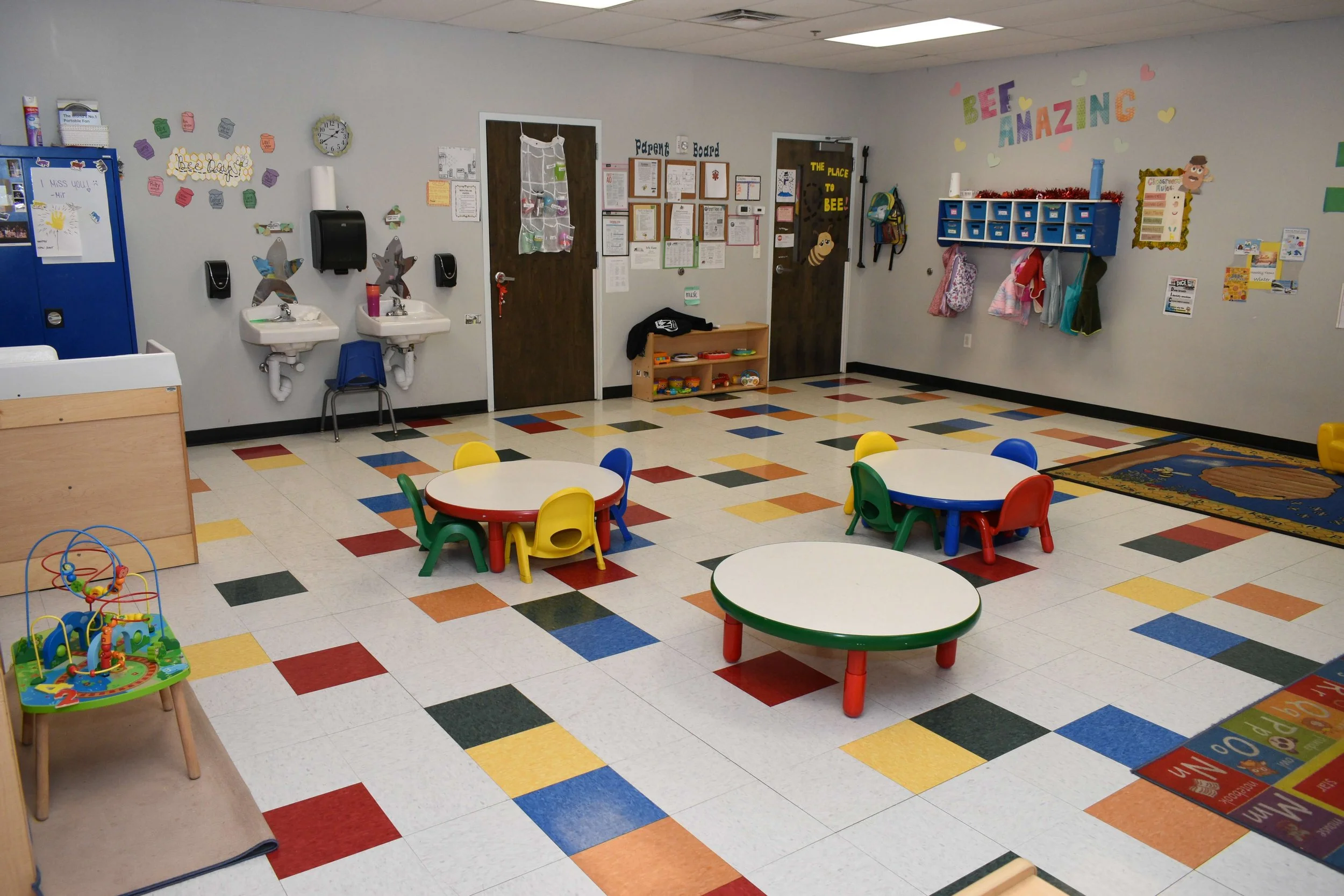 Colorful preschool classroom with small tables and chairs, a toy on a stand, and various decorations on the walls.