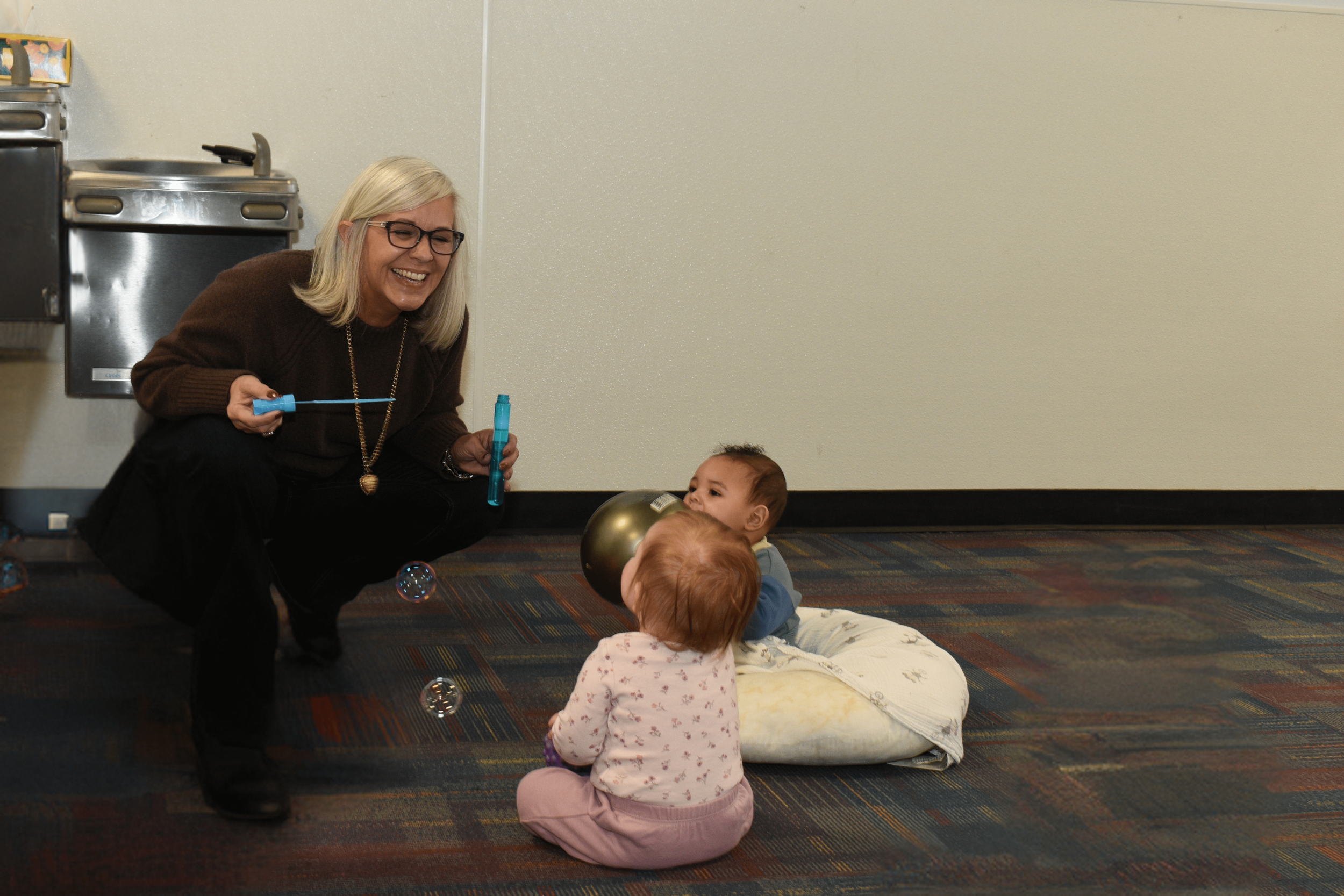 A woman playing with two young children on a carpeted floor in an indoor setting, with bubbles floating around.