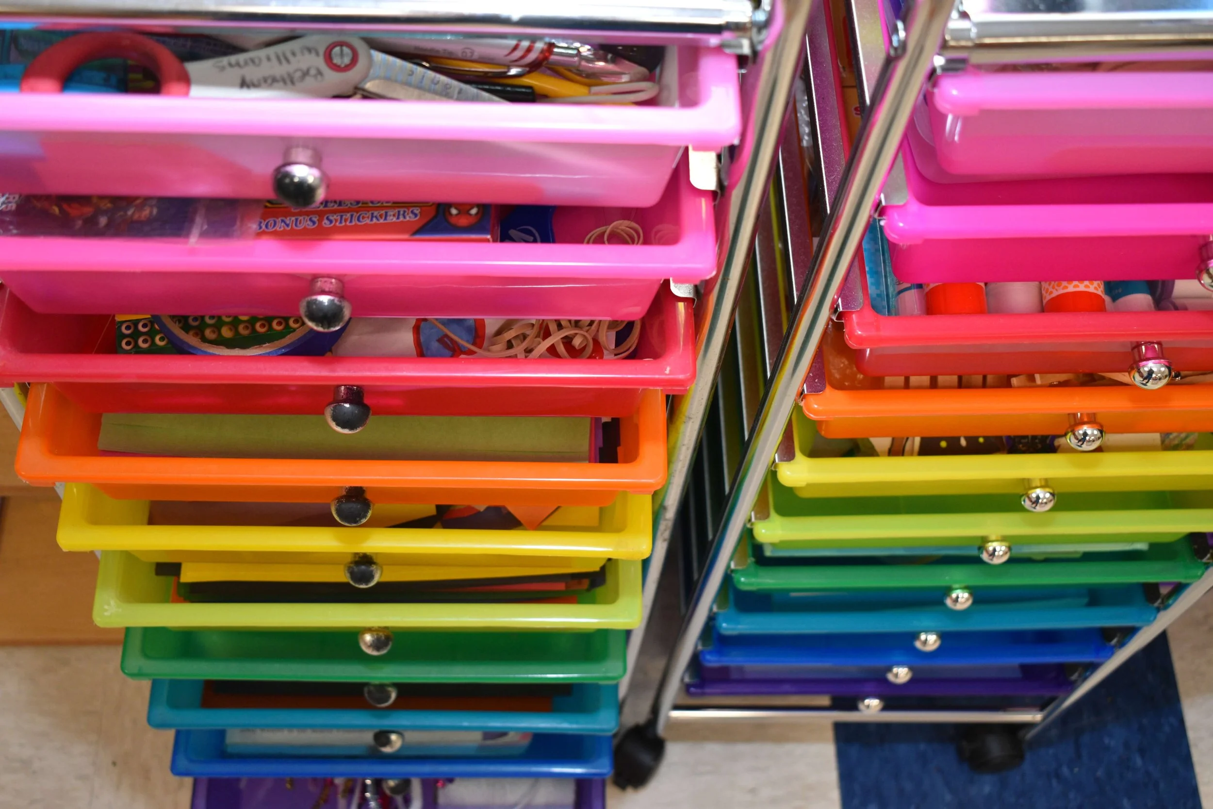 Colorful plastic drawers filled with office supplies such as pens, scissors, stickers, and rubber bands.