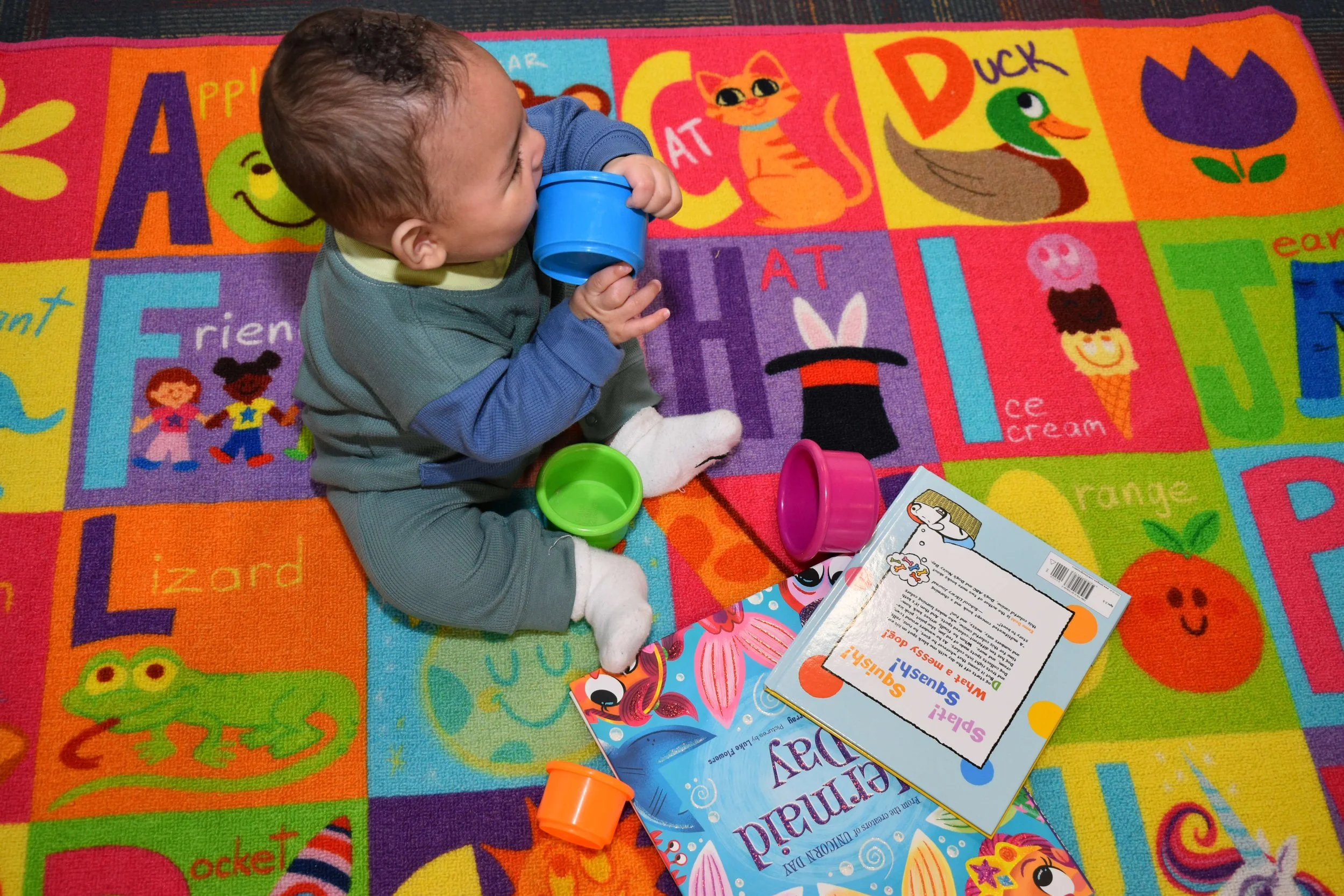 Young child sitting on a colorful alphabet-themed carpet, drinking from a blue cup, with various toys and books around him.