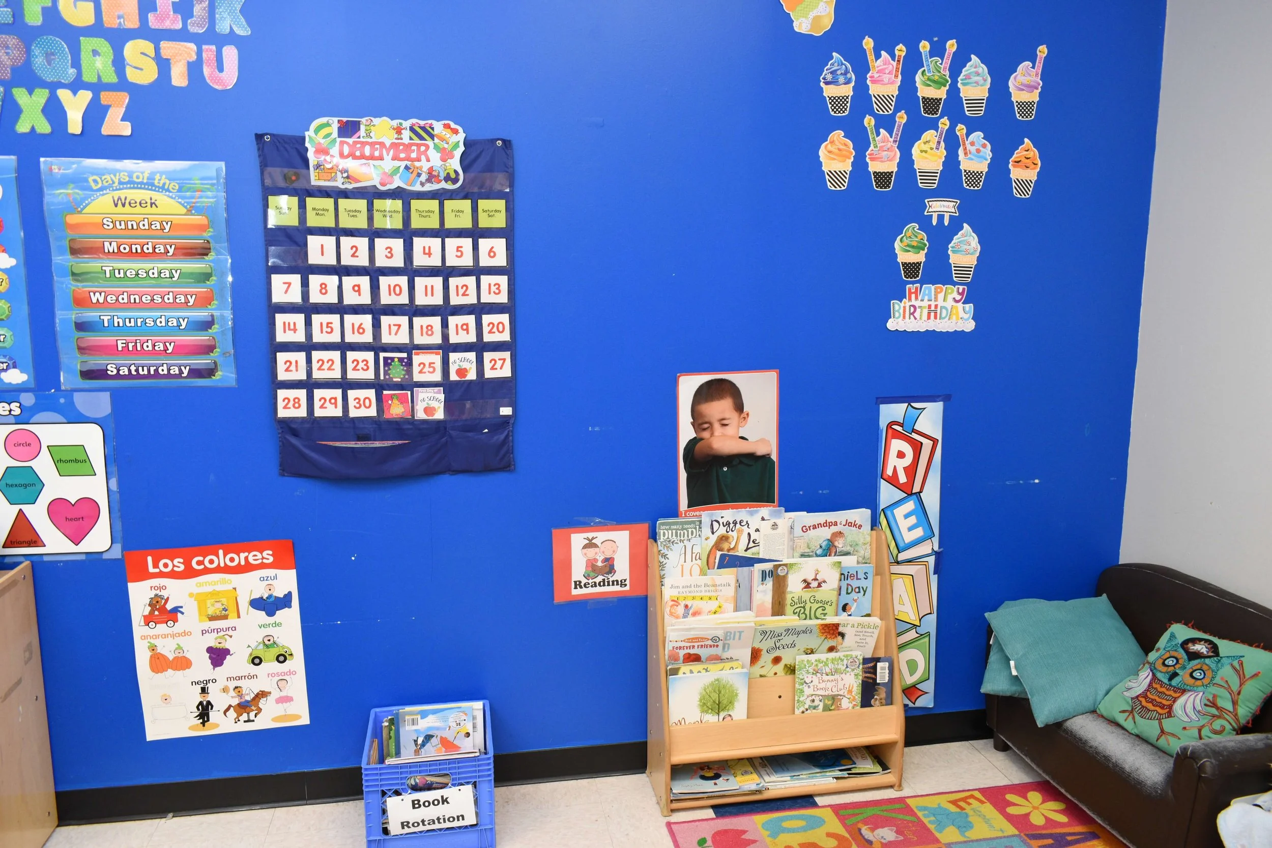 A vibrant preschool classroom corner with various educational posters and colorful decorations on a bright blue wall. There is a calendar for December, a weekly days of the week chart, and colorful ice cream cone charts. A small bookshelf holds child