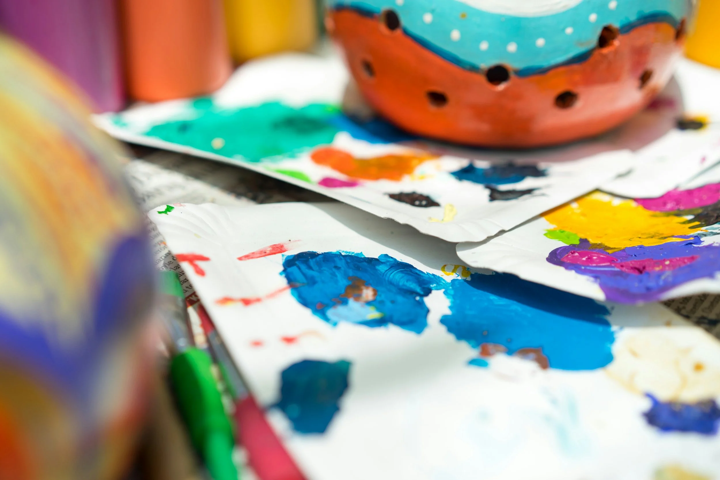 Close-up of a colorful painted ceramic bowl with various paint splatters and a partially visible paintbrush, surrounded by sheets of paper with different vibrant paint strokes.