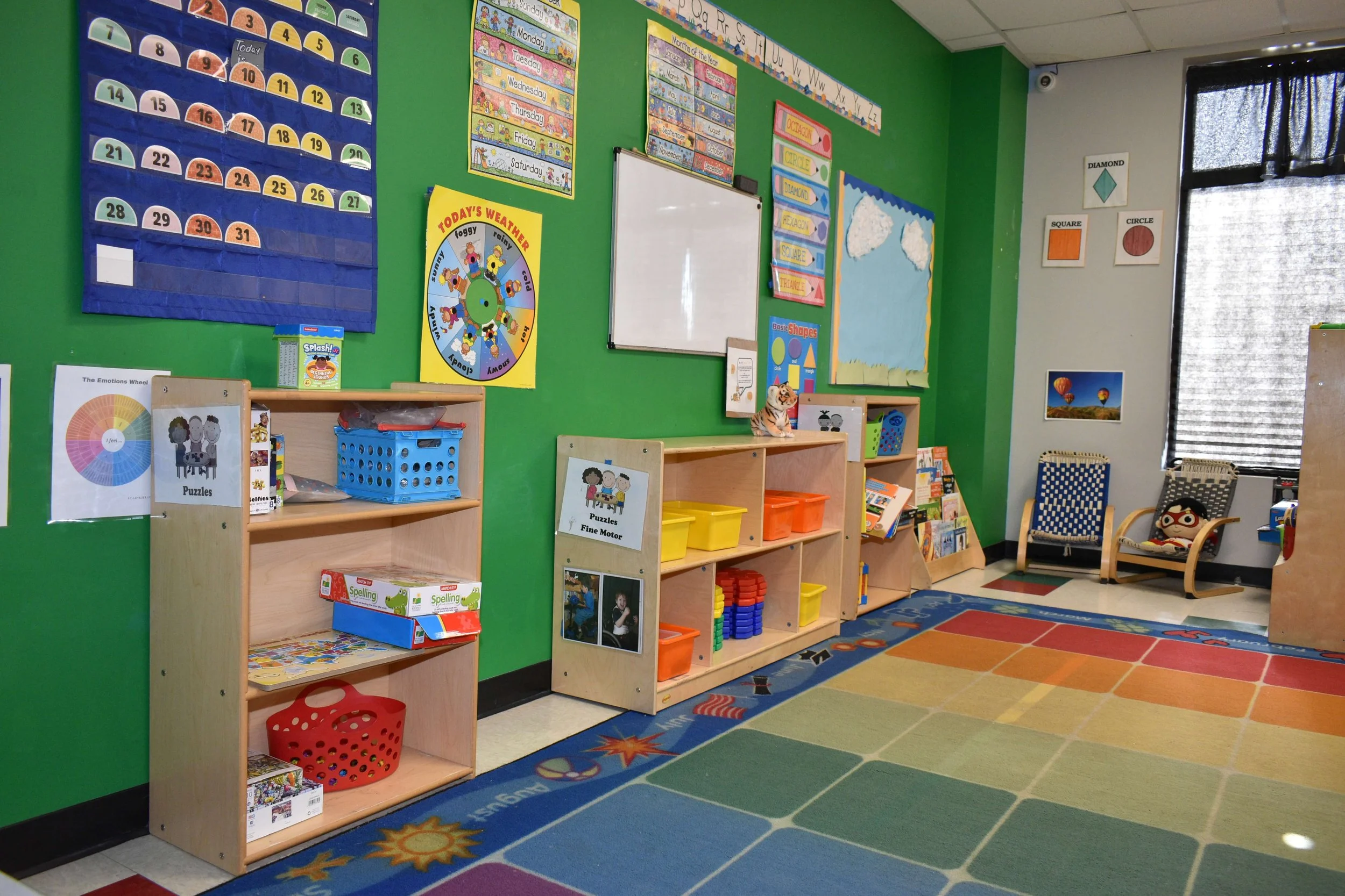 A colorful kindergarten classroom with educational posters, shelves filled with toys and books, and a vibrant area rug.