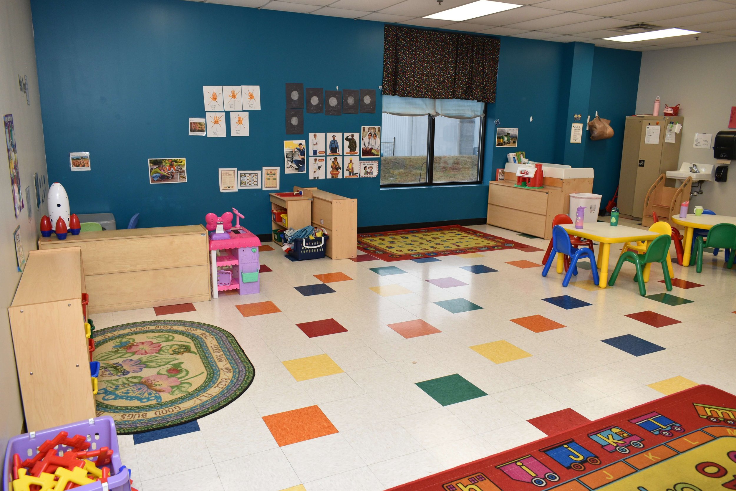 Brightly decorated preschool classroom with colorful tables, chairs, and educational toys, featuring wall posters, windows, and a patterned rug.