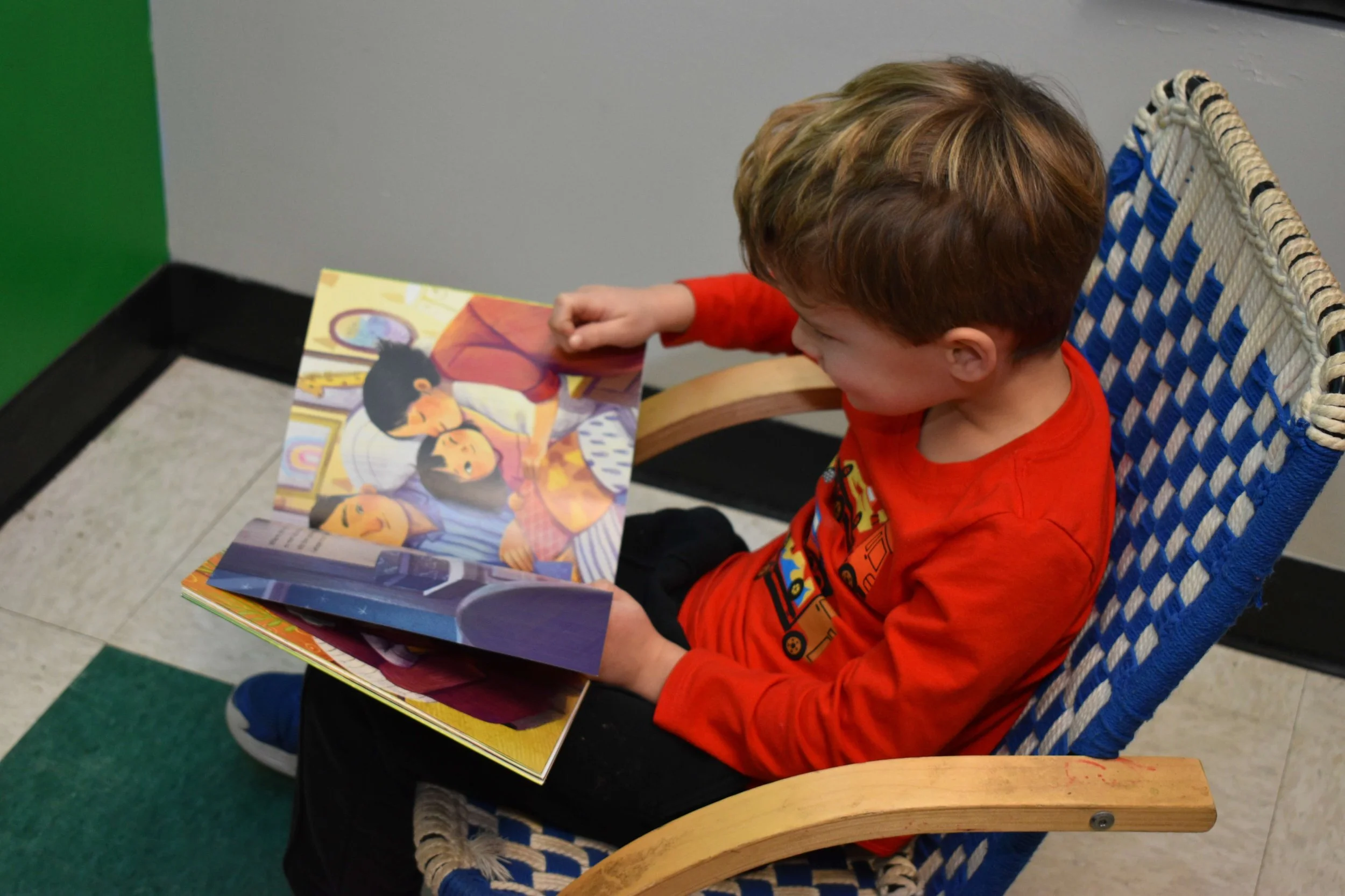 A young boy with light brown hair wearing a red shirt sitting in a blue and beige woven chair, reading a colorful children's book.
