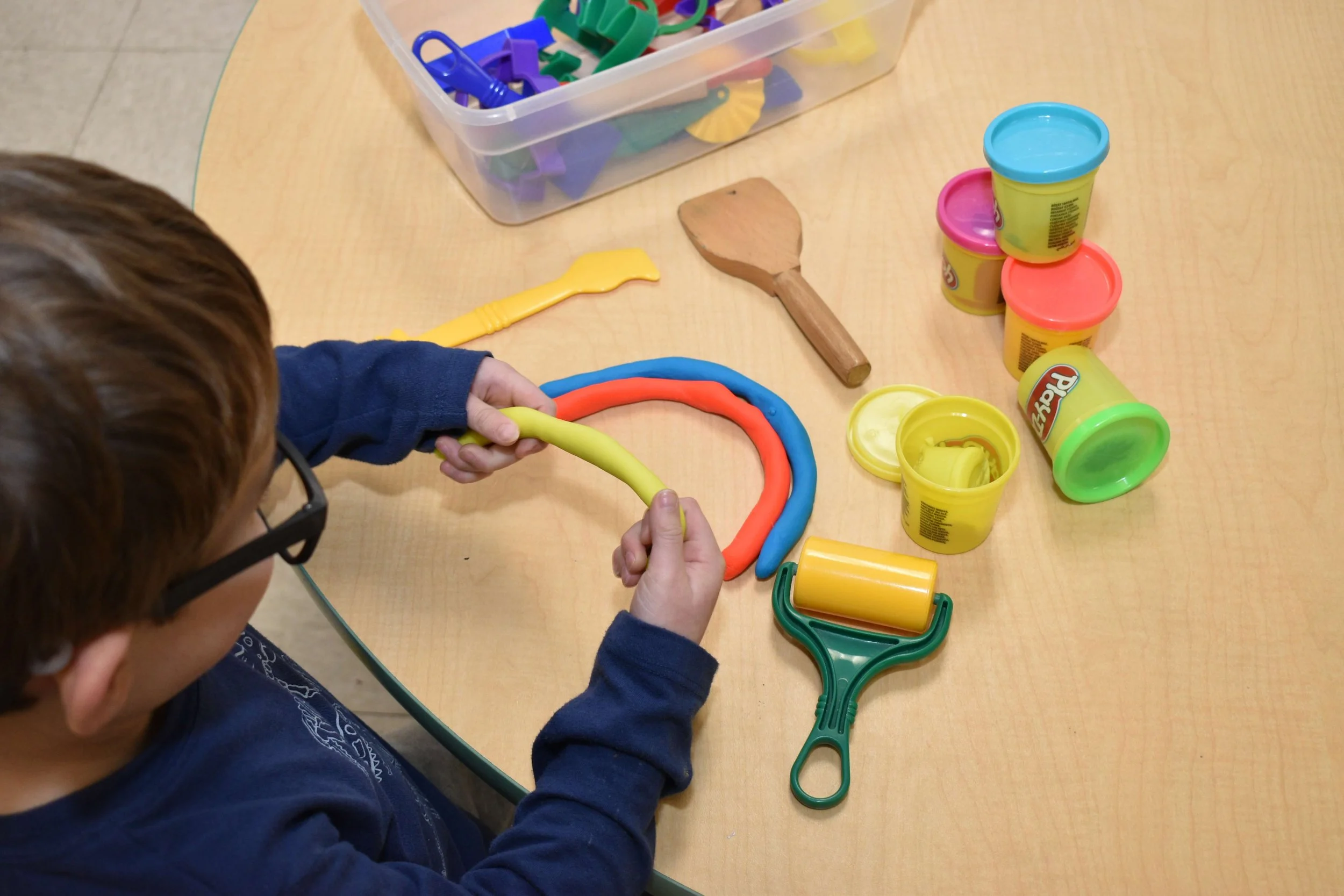 Child playing with colorful modeling clay and tools on a table, including a yellow roller, wooden spoon, and plastic knives, with a container of Play-Doh in various colors.