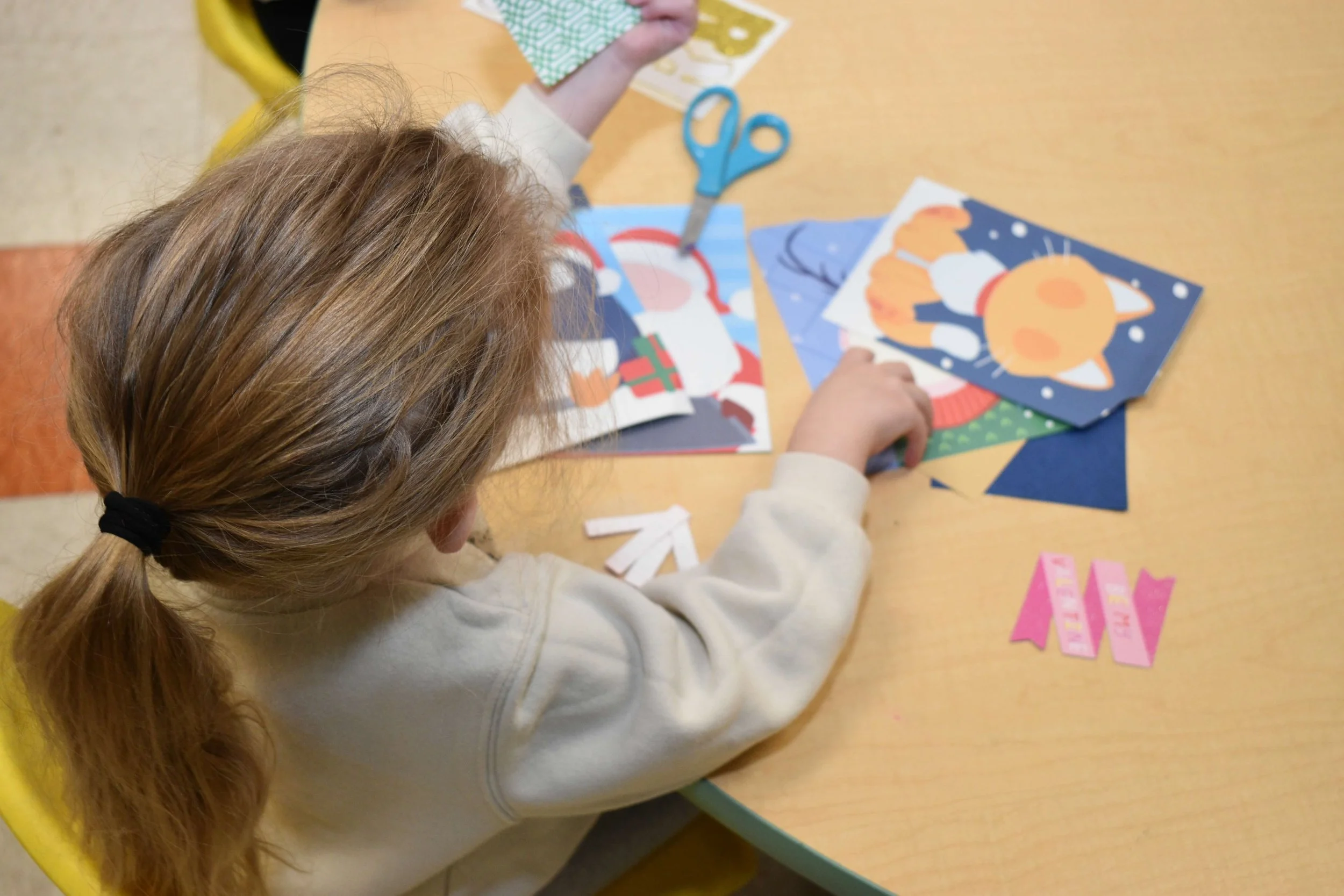 A young girl with red hair tied in a ponytail, wearing a light-colored sweatshirt, sits at a table working on a holiday craft project with colorful paper and scissors.