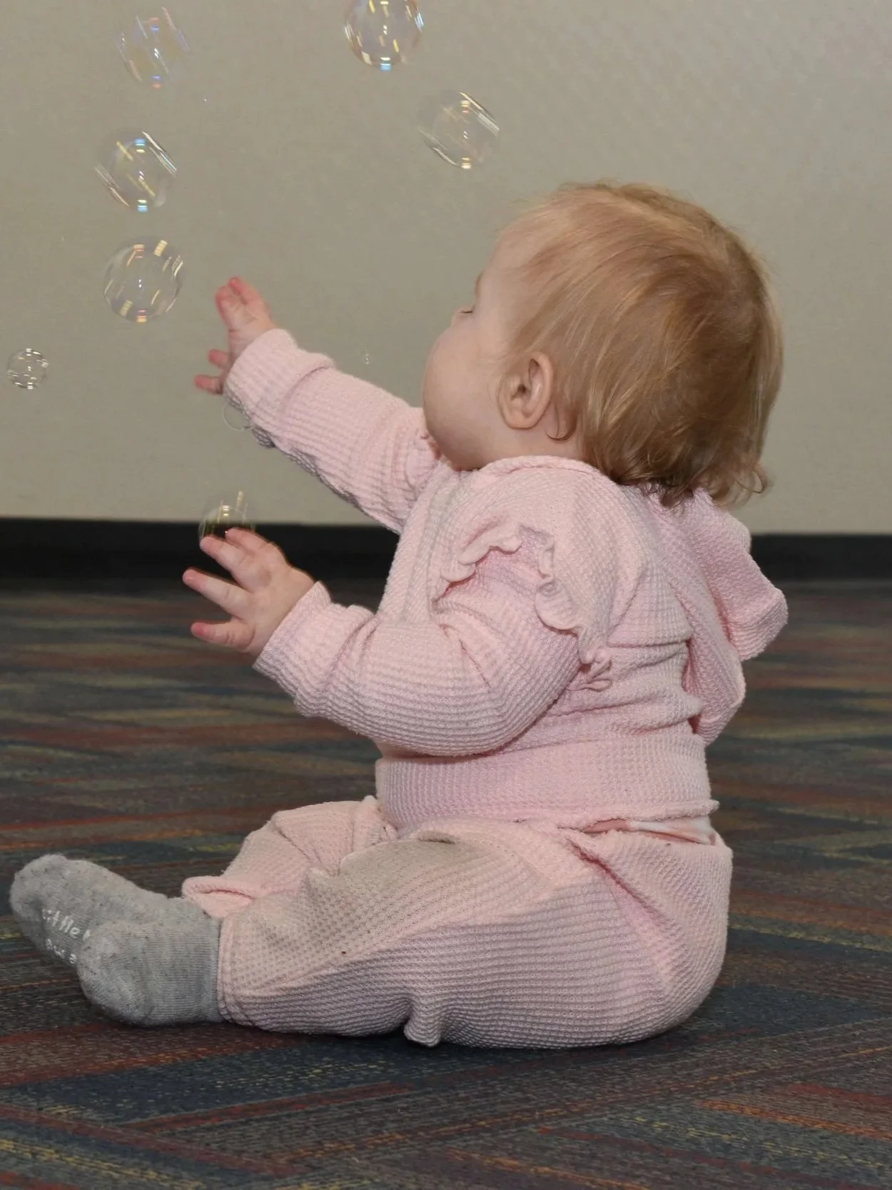 A young child with light brown hair, wearing a pink textured sweater and matching pink pants, is sitting on a multicolored carpet and reaching out to touch floating soap bubbles.