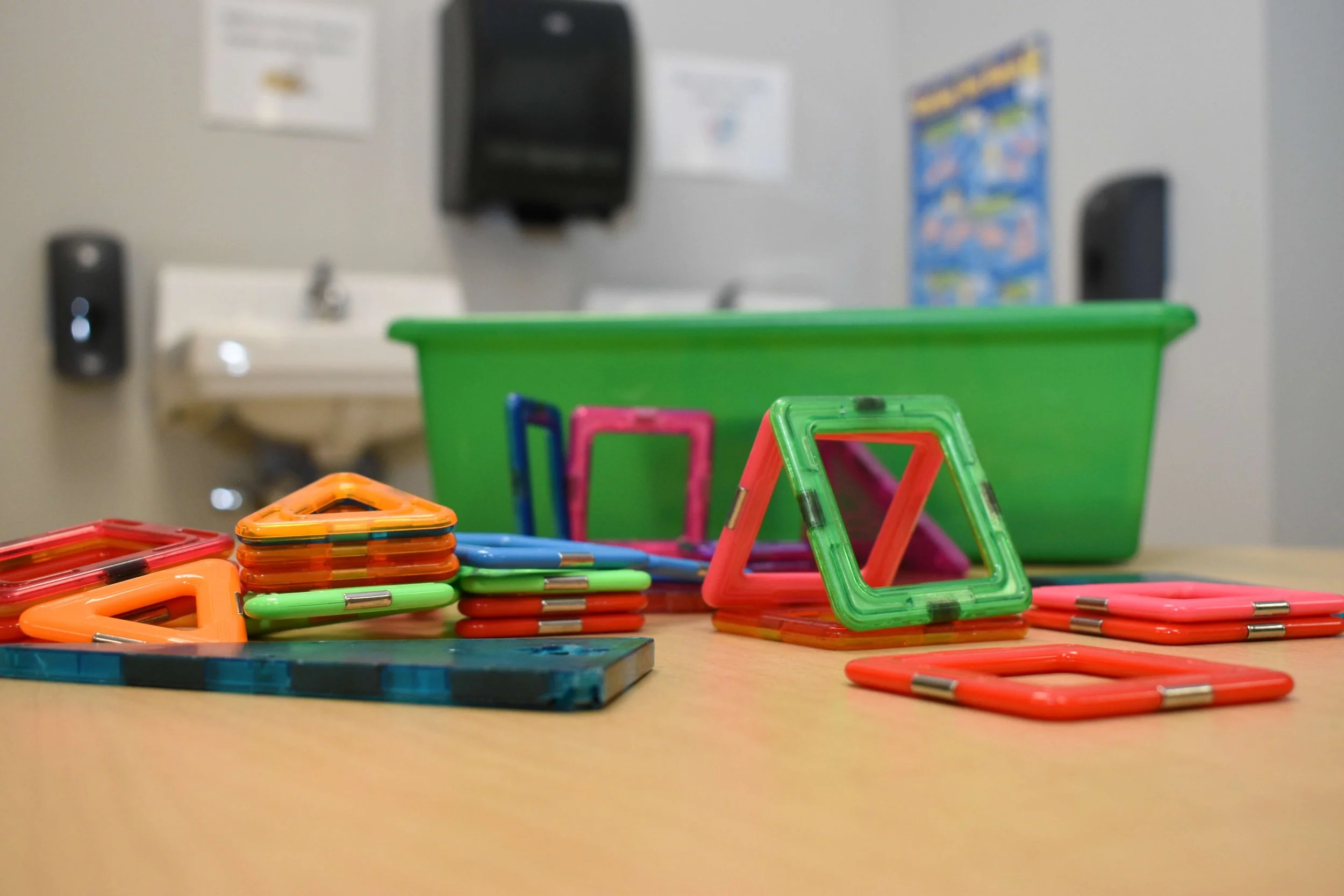 Colorful magnetic tiles arranged on a classroom table with a green storage bin behind them.