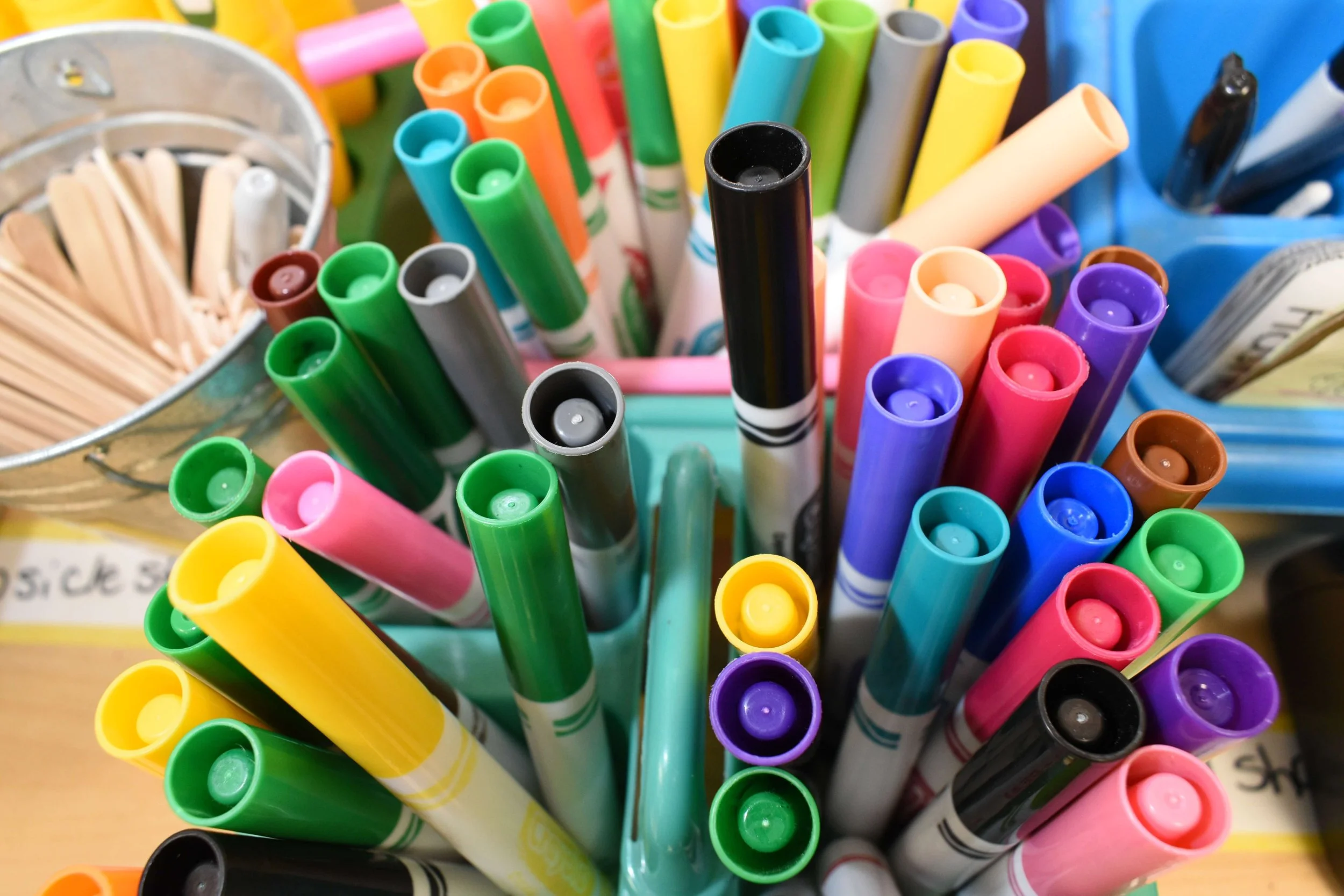 A collection of colorful markers in shades of blue, green, yellow, pink, purple, black, and orange arranged in a container.