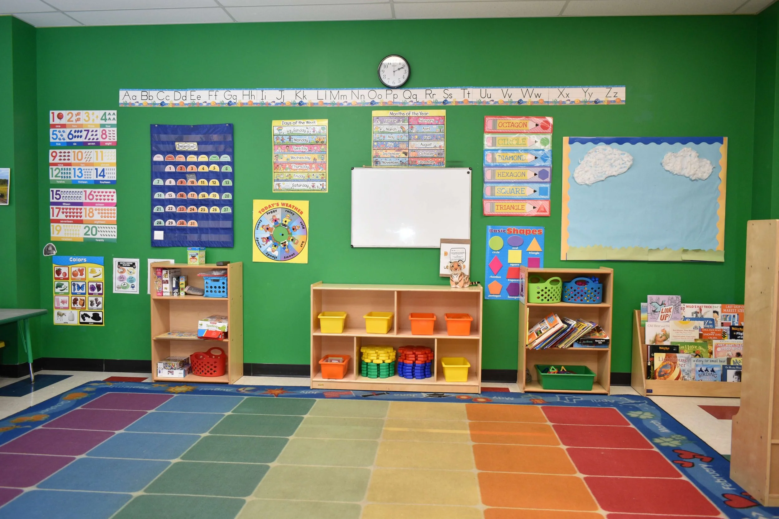 Colorful classroom with green walls, educational charts, and shelves filled with books and toys. The room has a soft, multicolored carpet and a weather wheel for learning activities.