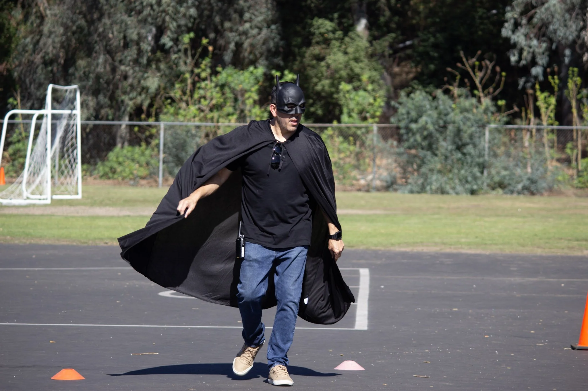 A man dressed as Batman running on an outdoor sports court with orange cones arranged around him.
