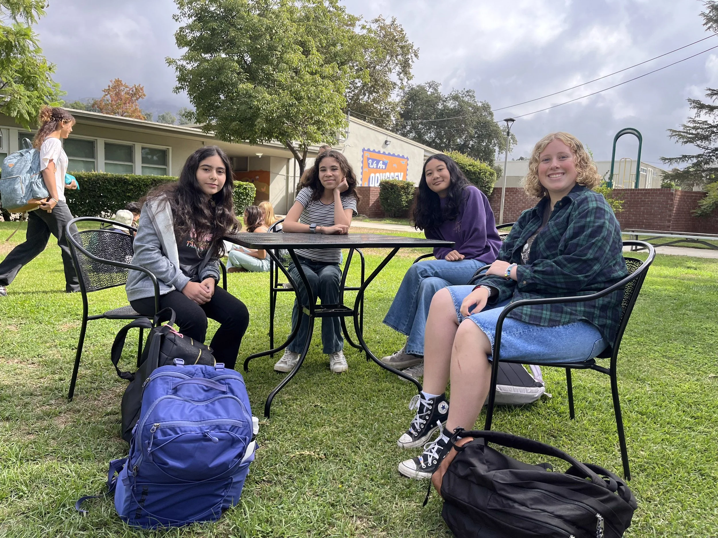 Four teenage girls sitting at a table on a grassy school yard, with backpacks on the ground, enjoying a break. One girl is walking past in the background. A school building is visible behind them.