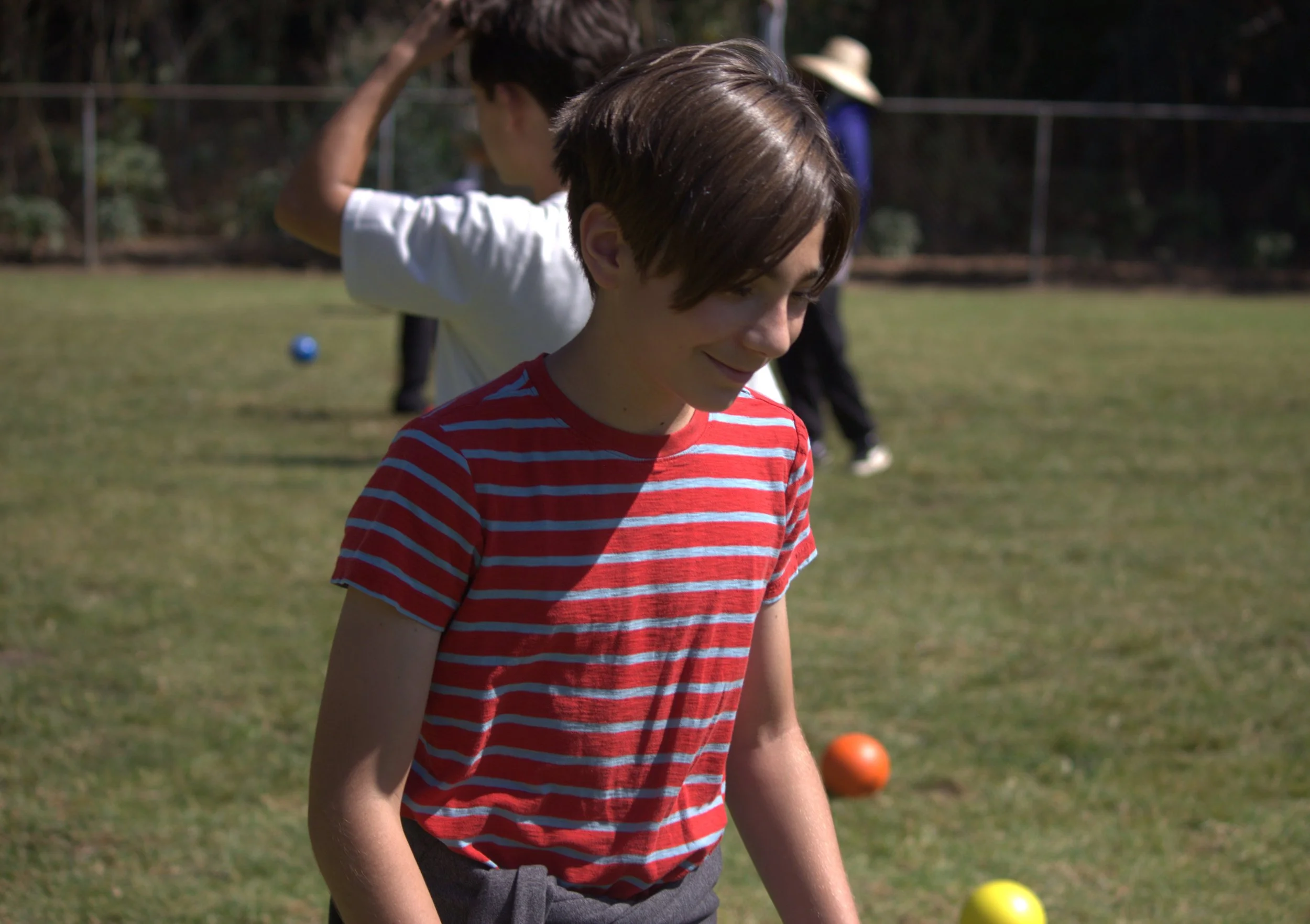 A boy with brown hair in a red and white striped t-shirt smiling outdoors on a grassy field with other children and a woman in the background.