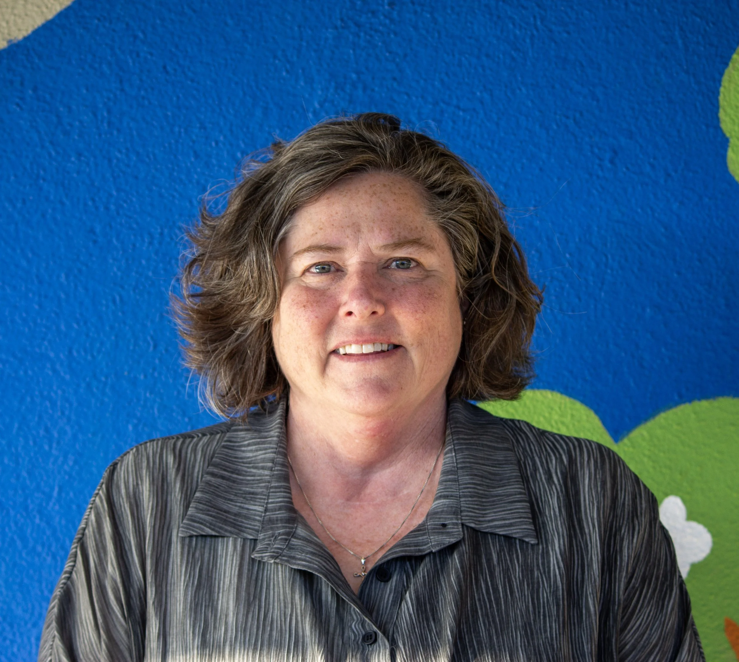 Close-up of a smiling woman with shoulder-length brown hair, wearing a dark top.